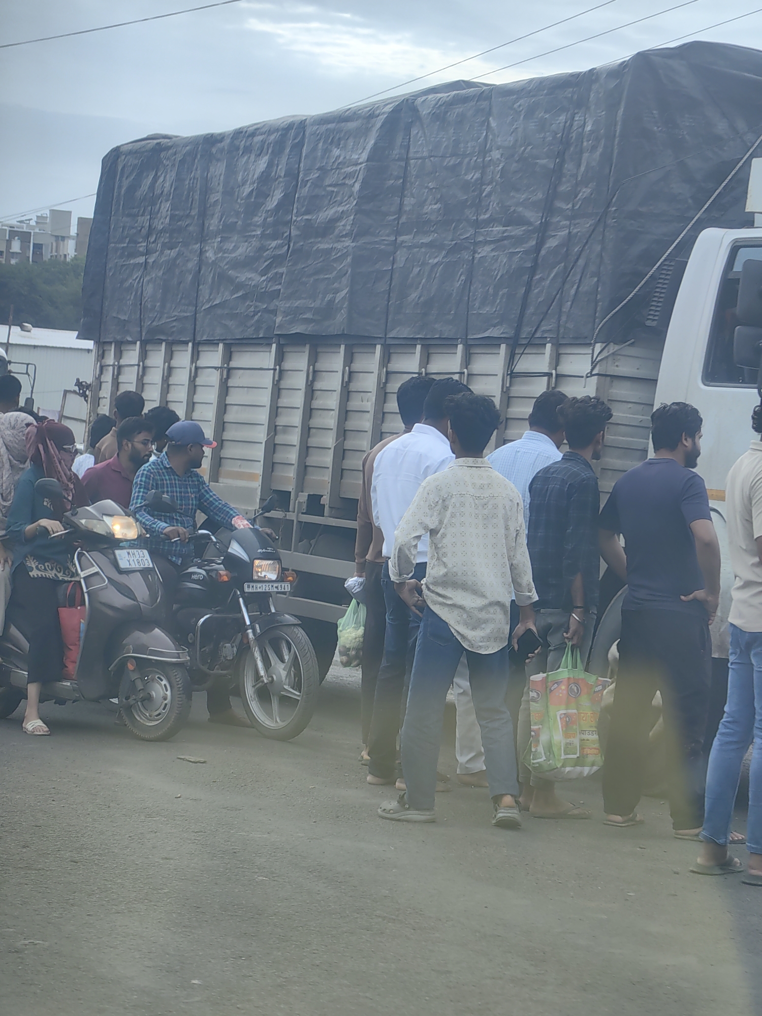 In Ranjangaon, India, a group of people are gathered on a dusty road beside a large, tarp-covered truck. Several individuals are standing near the truck's rear, some holding shopping bags. In the foreground, two-wheelers are visible, with a scooter and a motorcycle parked closely together. A man in a plaid shirt is seated on the motorcycle, while a woman in a headscarf is on the scooter. The scene suggests a busy roadside environment, possibly during a stop or a pause in transit. The sky is overcast, and the general ambiance is one of everyday life unfolding on an Indian road.
