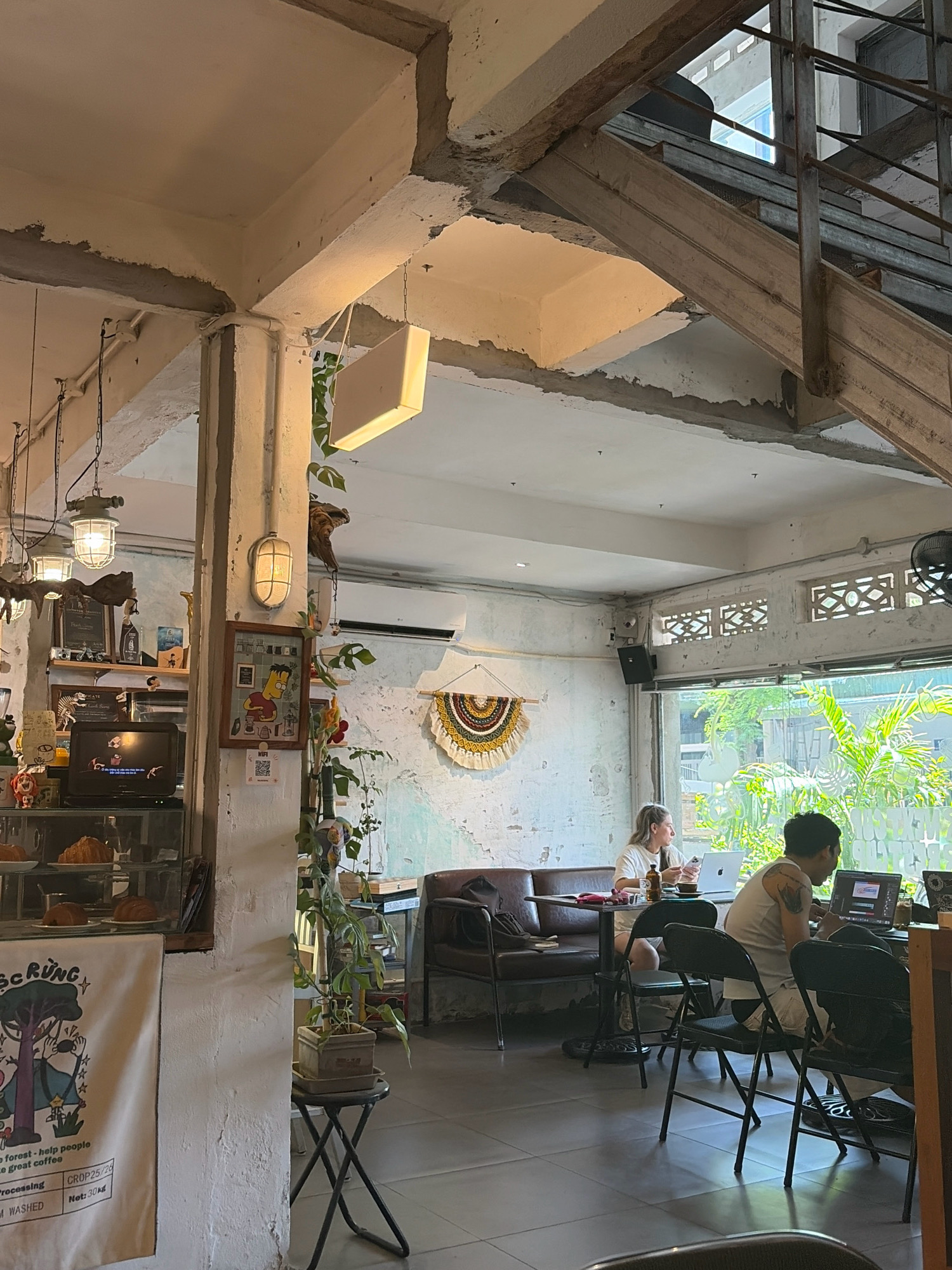 The image depicts the interior of a cafe in Da Nang, Vietnam. The cafe has an industrial-chic aesthetic with exposed concrete beams and rough-textured walls. Natural light streams in from a large window, illuminating the space and highlighting the lush greenery outside.
Two individuals are present in the cafe, both seated at tables and engaged with their laptops. One person, with blonde hair, is wearing a white t-shirt and appears to be working on a laptop. The other person, with dark hair and tattoos on their arm, is wearing a white tank top and also appears to be focused on their laptop.
The cafe is furnished with dark leather sofas and chairs, creating a comfortable seating area. Decorative elements include hanging plants, wall art, and vintage-style lamps, contributing to the relaxed and bohemian atmosphere. A display case near the front showcases pastries, and a banner with illustrations hangs on the wall, suggesting the cafe's branding or a local event. The flooring is tiled. The overall impression is that of a contemporary cafe offering a workspace environment.