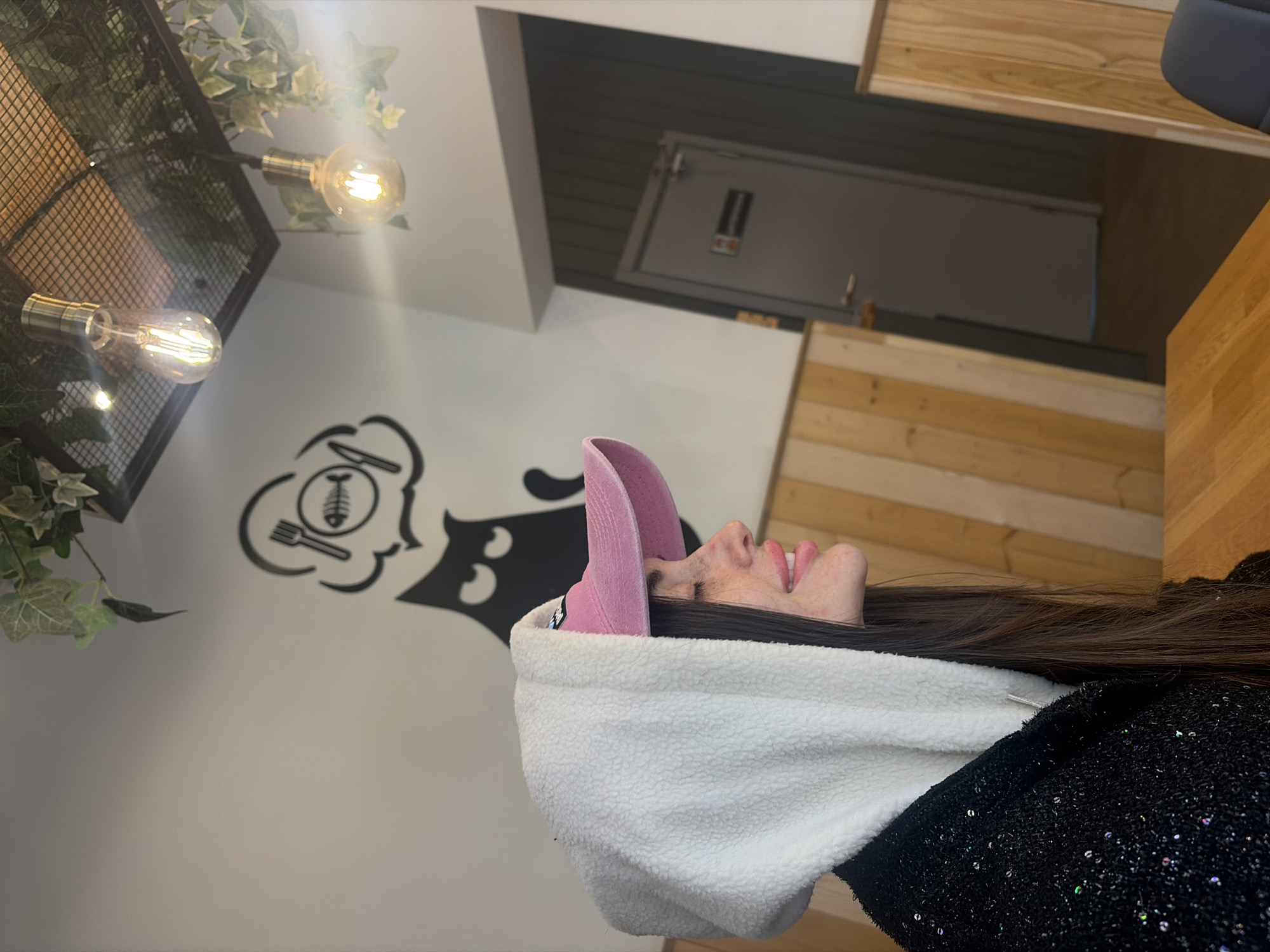 A woman wearing a white hooded sweatshirt and a pink baseball cap is indoors. Her face is visible in profile. The sweatshirt covers her hair which is brown. She is wearing a sequined black top. The background includes a white wall with a black decal of a cartoon-style chef's head with a fish skeleton, fork, and knife. There is a light fixture with two Edison bulbs. Some green plants are hanging from the fixture. The top of a door is in the background, as well as some wood paneling. The scene does not appear to be located in Saint Petersburg, Russia.