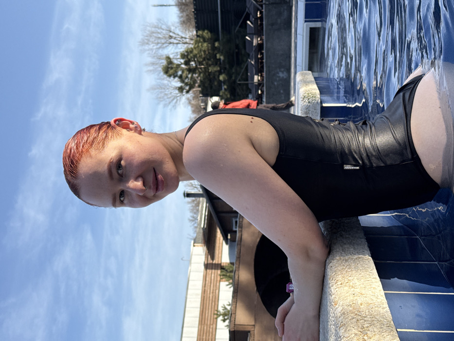 The image presents an outdoor scene at **Термы "Лес и Река" (Therms "Forest and River")** in Rostov-on-Don, Russia, on a clear, sunny day.
The primary subject is a light-skinned adult female, positioned from the mid-torso upward, submerged in a body of water. She has reddish-orange, wet hair and multiple piercings on her left ear. Her forearms are resting on the light-colored, textured coping of the pool edge. She is wearing a black, one-piece swimsuit, which has a subtly shiny texture on the lower torso and a laced detail on the left lateral torso. Water droplets are visible on her exposed skin, including her shoulders and right arm. She is looking directly at the camera with a slight smile.
The environment includes a blue-tiled outdoor swimming pool. In the background, a rustic-style brown building with a distinctive arched, bridge-like structure is visible. Adjacent to this, another building features horizontal light-colored paneling, with a dark vertical pipe (possibly a chimney) extending upwards. Various trees, some deciduous with bare branches and others coniferous, are scattered across the background. To the right, an area with dark lounge chairs is visible, and an indistinct individual wearing a red jacket is seen walking away from the camera. The sky is clear blue with sparse white clouds.