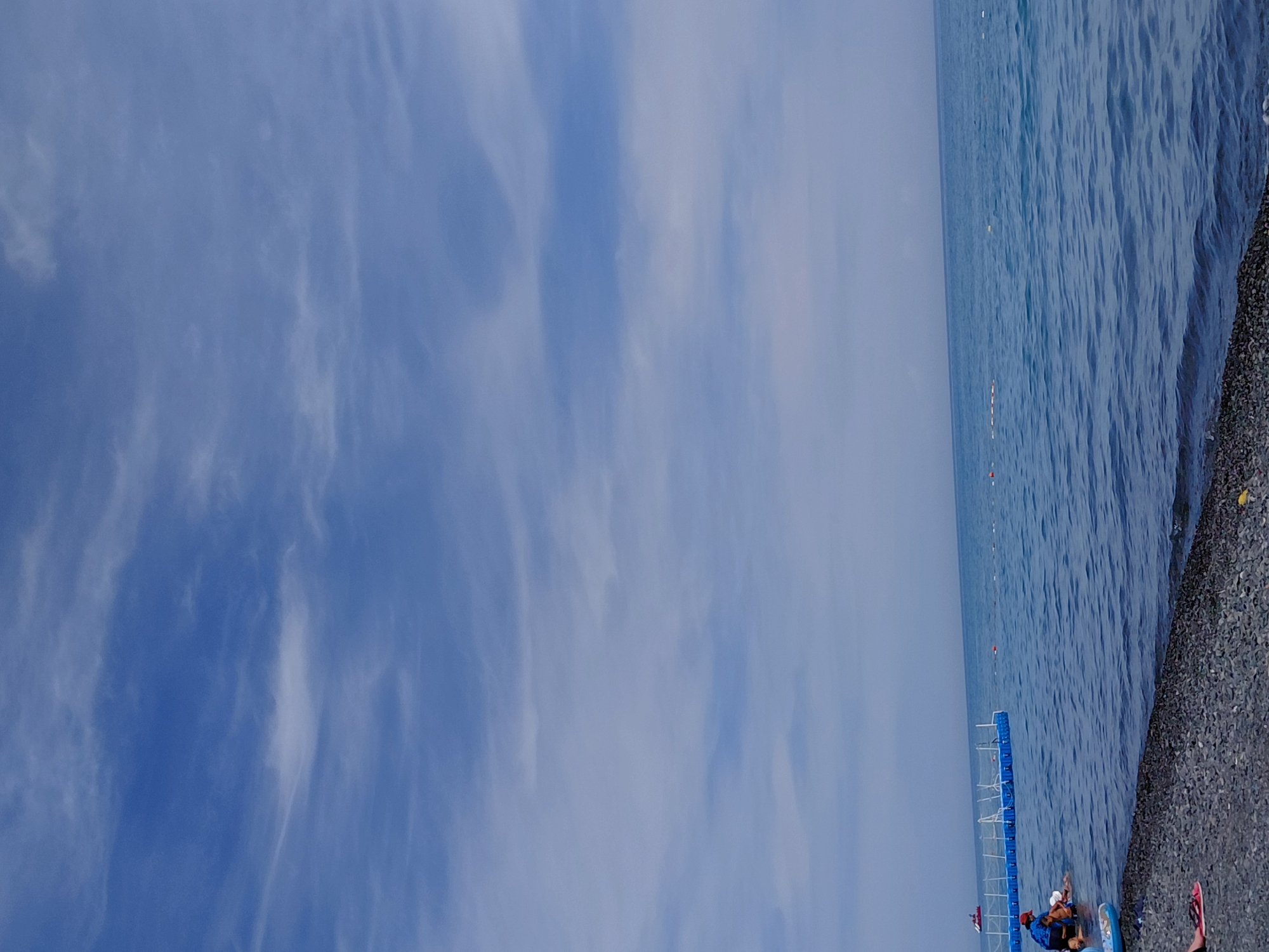 The image depicts a beach scene with the sky taking up the majority of the frame. The sky is a bright blue with scattered white clouds, some appearing wispy and others more defined. In the distance, the horizon line separates the sky from the sea, which is a deep blue and slightly choppy.
On the left side of the image, closer to the viewer, is a pebble beach. Several people are visible near the water's edge. One person, wearing a blue shirt and a red hat, appears to be interacting with two children in the shallow water. A blue floating structure, possibly a walkway or barrier, extends into the sea from the left. A small red boat is visible further out in the water.
The foreground is dominated by the dark pebbles of the beach. A pair of red flip-flops are visible on the lower left corner of the image. The overall impression is a typical summer day at the seaside. The location is identified as Ақши, Kazakhstan.
