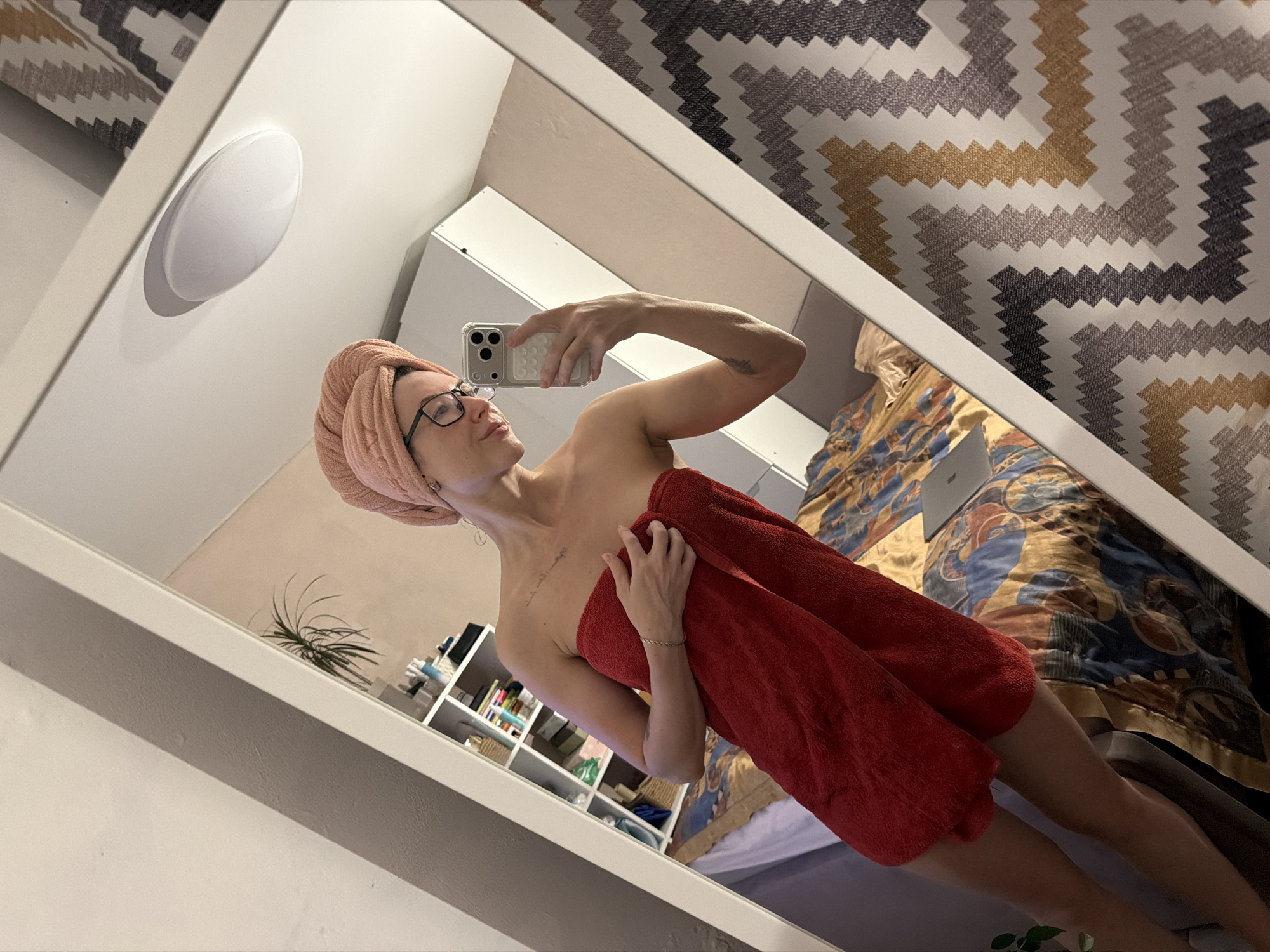 A female subject is depicted in a mirror selfie, standing indoors. Her hair is wrapped in a peach-colored towel, and her body is covered by a red towel, extending from her chest to her upper thighs. She wears dark-framed eyeglasses and holds a light-colored smartphone with a triple-lens camera in her right hand, positioning it to capture her reflection. Her left hand is stabilizing the red towel. A faint linear tattoo is visible on her upper left chest/shoulder area.
The reflection reveals a domestic interior. To the left, a white shelving unit is stocked with various household items, including baskets and bottles. A green potted plant rests on the top shelf. A bed with a patterned duvet, featuring blue, gold, and dark red abstract shapes, is visible in the lower center and right portions of the reflection, with a silver-colored laptop positioned open on it. Another portion of a bed with a plain blue sheet is visible further left. The wall behind the shelving appears to be a light, solid color. To the right of the subject, a wall or curtain exhibits a geometric zig-zag pattern in shades of white, grey, beige, and yellow. A white, circular flush-mount ceiling light fixture is visible in the upper right quadrant of the reflection. The overall environment suggests a private residence, likely a bedroom.
The specific location within the city of Perm, Russia, cannot be determined from the visual evidence presented in the image.