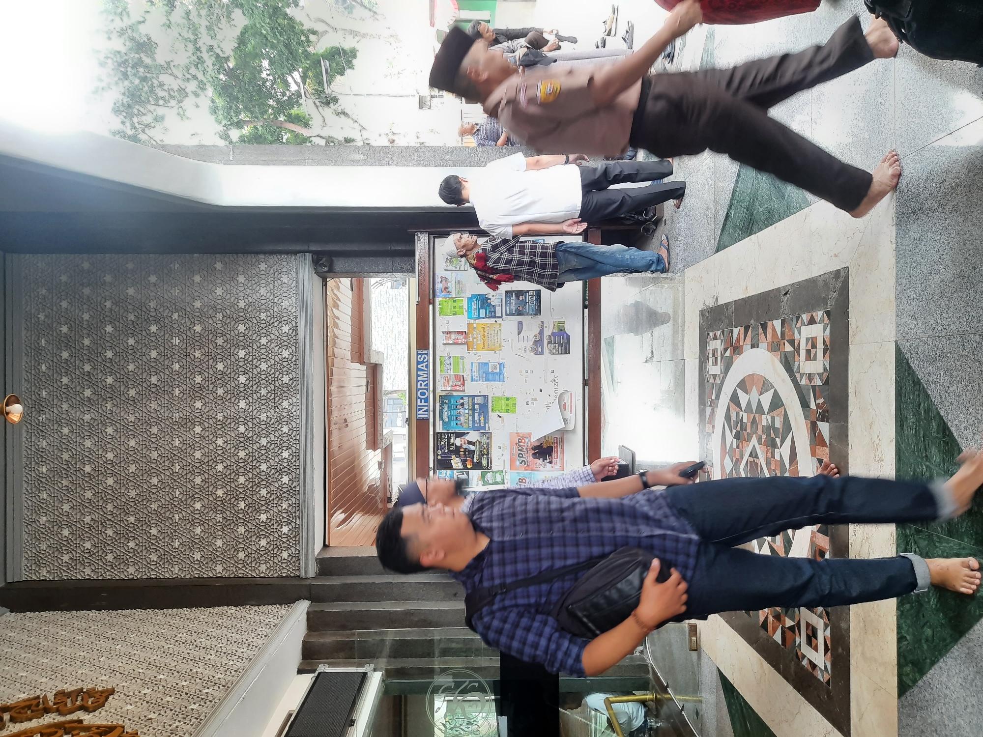 The image captures a scene at the entrance of a building, likely a mosque or community center, with a prominent information board displayed on the wall. Several individuals are visible, some walking and others standing. In the foreground, a man wearing a blue checkered shirt and dark jeans walks towards the right, carrying a black bag slung across his chest. His left foot is bare, and his right pant leg is rolled up. To his left, another man with a similar shirt and jeans is partially visible.
Behind them, an information board is covered with various posters and flyers. Among them, one poster features an image of a man in white and text in Arabic and Indonesian, possibly an announcement or religious information. Another poster, labeled "SPMB," also displays text and a phone number.
Further back, a group of people are seen near the entrance. One man, wearing a white shirt and dark pants, stands facing away from the camera. An elderly man with a white cap and a red and white scarf is also visible near the information board. To the right, a person in a brown uniform with insignia on the chest is walking, their face blurred due to motion. Other individuals are also present, some with covered heads, suggesting a religious context.
The environment appears to be an outdoor or semi-outdoor space, with trees visible in the background through an archway. The floor is tiled with an intricate geometric pattern, featuring shades of green, beige, and brown. The architecture includes textured walls and columns. The lighting suggests it is daytime, with some bright light coming from the right side of the frame. The scene appears to be taking place in Bandung City, Indonesia, given the visual context of the information displayed on the board and the common architectural styles.