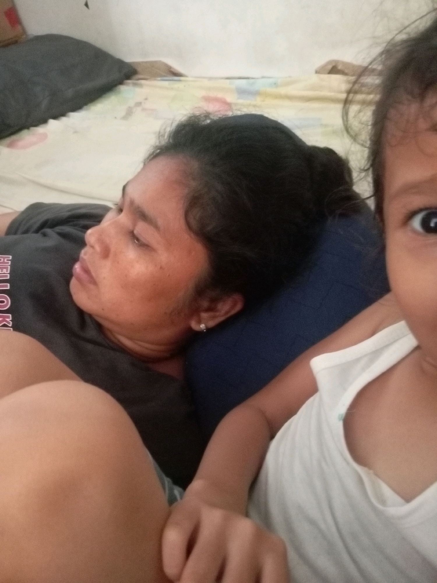 The image shows a close-up view of two individuals lying down, possibly on a bed. The person in the foreground on the right is a young child, whose face is partially visible, looking directly at the camera. They are wearing a white tank top. The person in the background on the left is an adult, who appears to be resting or sleeping, with their head on a dark blue pillow. They are wearing a dark gray t-shirt with white lettering that is partially visible. The environment appears to be indoors, with a light-colored patterned bedsheet and a dark pillow visible. There is no discernible location within the city of Batam, Indonesia indicated in the image.