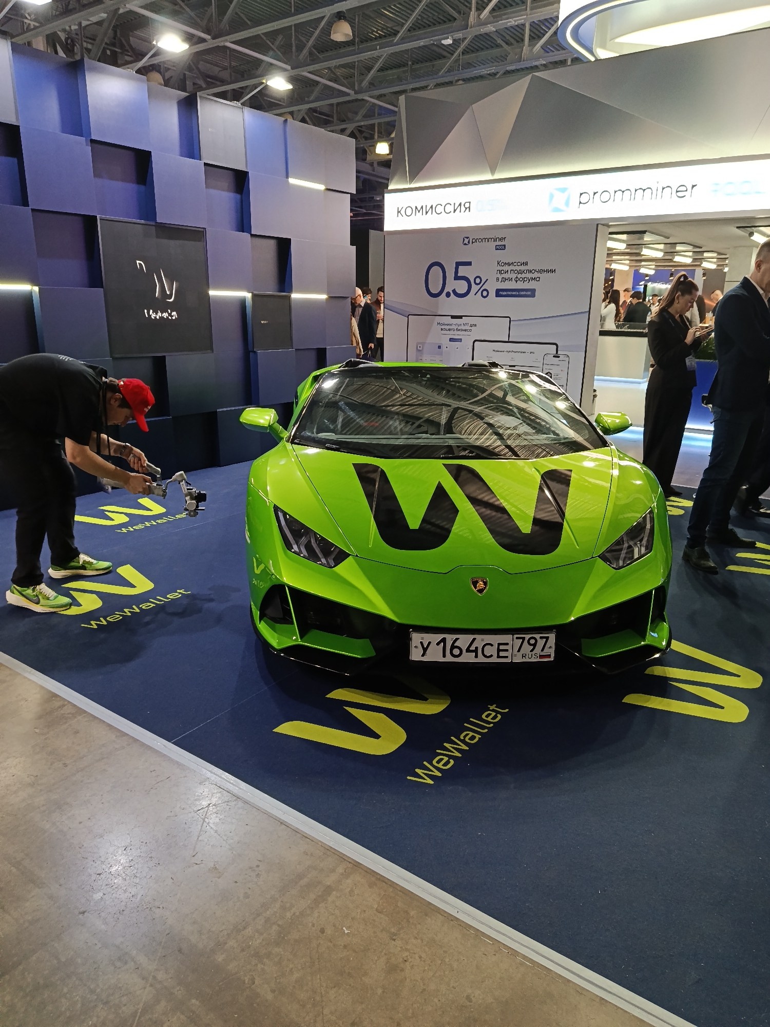 Here is a forensic description of the image:
The image depicts a bright green Lamborghini car displayed at an event. The car has the "WeWallet" logo on its hood, and the license plate reads "Y 164 CE 797 RUS". The car is positioned on a blue floor covering with a yellow "WeWallet" logo.
A man wearing a black shirt, black pants, and a red cap is standing to the left of the car, holding video equipment (likely a camera stabilizer). He appears to be filming or photographing the car.
In the background, there is a blue wall design and a Promminer booth advertising a 0.5% commission. Several people are present near the Promminer booth, including a woman in a black dress and a man in a suit.
Based on the license plate and textual information, the location is likely within Russia. The OCR identified text includes “Khimki” on the license plate, pointing to the city of Khimki in Russia as the potential location.