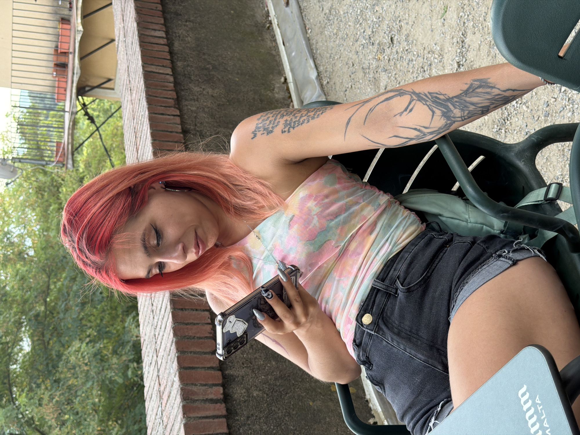 A young woman with bright pink hair sits in an outdoor cafe, looking down at her phone. She is wearing a tie-dye one-shoulder top and denim shorts. Her left arm is adorned with a large tattoo of a deer's head. The cafe appears to be on a slight incline, with a brick wall and greenery in the background, suggesting it could be located in Hostalric, Spain. The overall scene depicts a casual moment of a person engaged with their device in an outdoor setting.