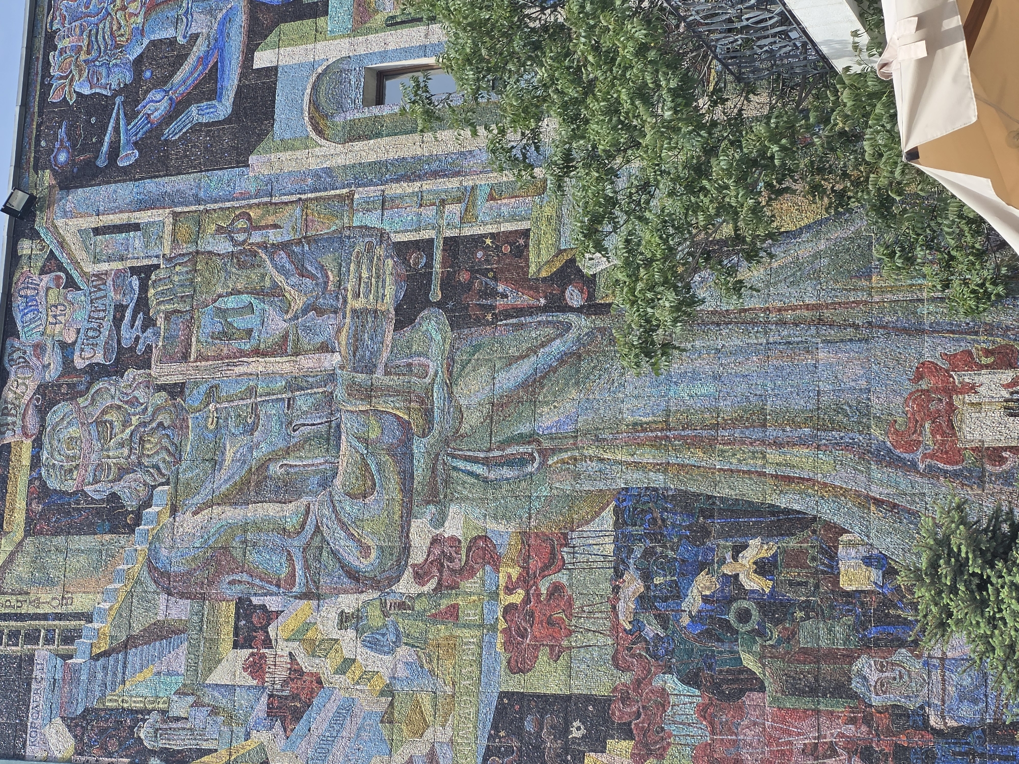 The image depicts a large, colorful mosaic mural on the side of a building. The mural is in Krasnodar, Russia, featuring a stylized depiction of a person seated, holding hands, with geometric patterns and symbolic imagery throughout. The mosaic uses a wide range of colors, including blues, greens, reds, and browns. Parts of the mural are obscured by a tree and an awning. There is a window visible in the mural. The scene is a daytime exterior shot.