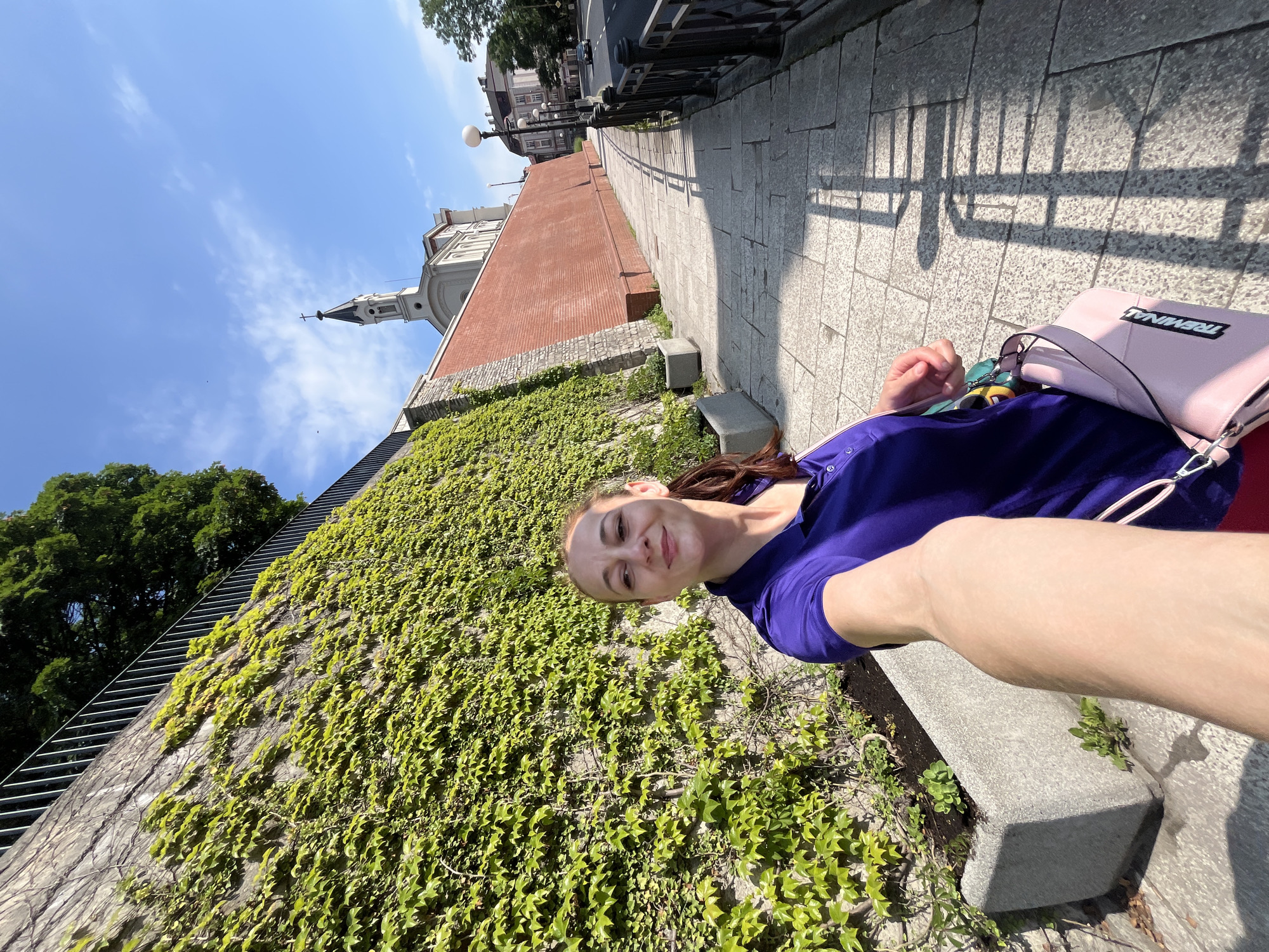 The image captures a woman taking a selfie as she walks along a sidewalk in an urban environment. She is wearing a purple polo shirt and a pink handbag is slung over her shoulder. To her left is a steep, stone wall covered in lush green ivy, and to her right is a paved sidewalk that leads towards a street with buildings in the background. The sky is clear and blue with some scattered clouds. The scene appears to be from a sunny day, with shadows cast by a railing along the sidewalk. In the distance, a church with a steeple can be seen. The overall impression is of a pleasant walk through a European-style city.