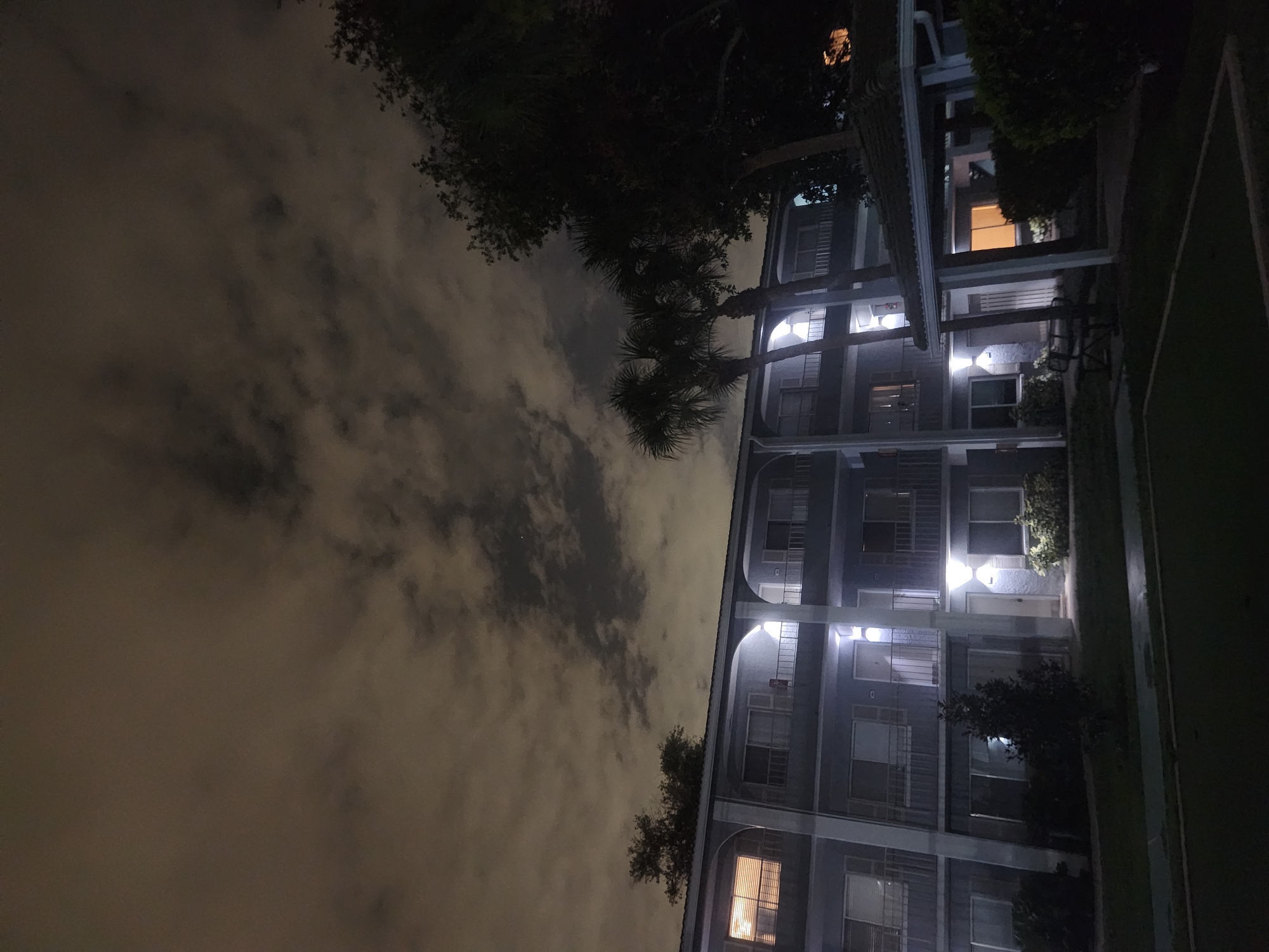 The image captures a nighttime view of a multi-story apartment building in Altamonte Springs, Florida, United States of America. The building is grey with white trim and features balconies on each level. Outdoor lights illuminate the balconies and doorways, creating bright pools of light. Palm trees are visible in front of the building, and a covered patio area with a bench is situated in the foreground. The sky above is cloudy, with the clouds illuminated by an unseen light source. The overall scene suggests a residential complex at night.