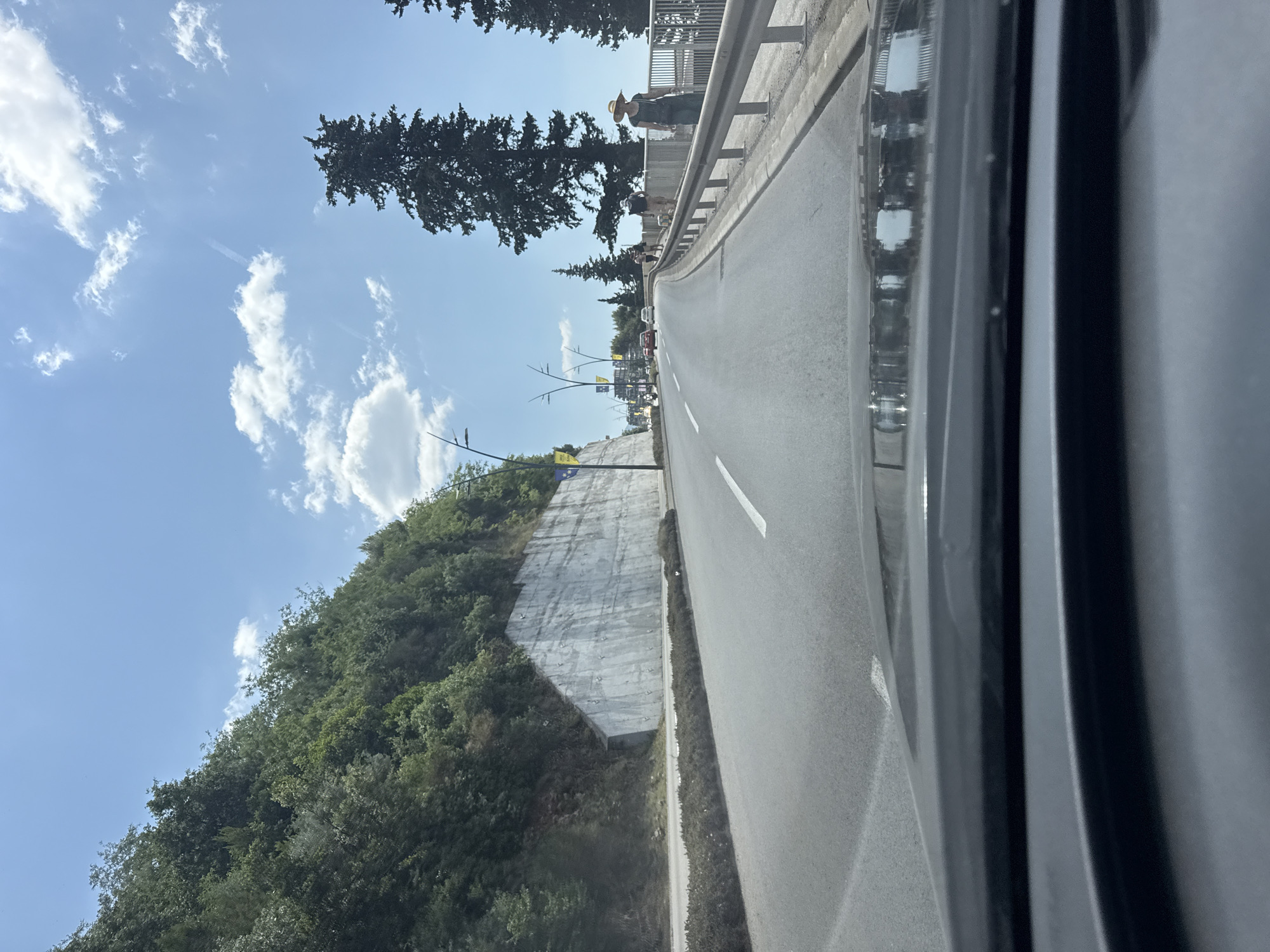 This is a first-person view from a vehicle driving on a road in Budva, Montenegro. The road is paved and has lane markings. To the left, a steep, tree-covered hillside rises, with a concrete retaining wall at its base. To the right, there is a sidewalk with a metal railing and several people walking. Tall coniferous trees are visible on the right side of the road. The sky is blue with scattered white clouds. There are other vehicles on the road in the distance. The foreground shows the dashboard and windshield of the vehicle.
