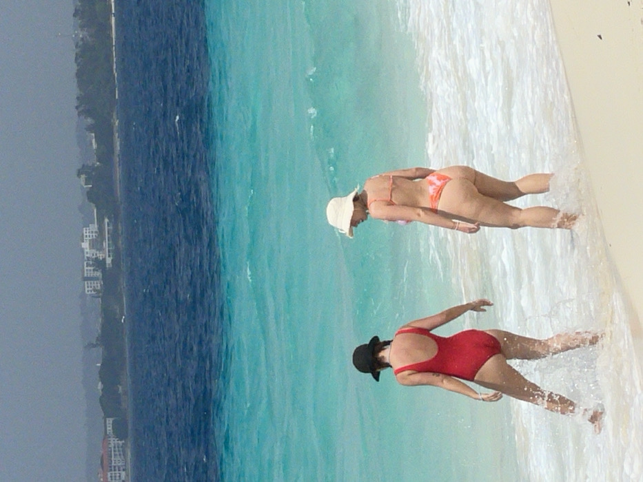 The image displays a daytime beach scene located in Zanzibar, Tanzania. Two adult females are partially submerged in the shallow, clear turquoise ocean water near a sandy shoreline. The individual on the left, viewed from behind, wears a red one-piece swimsuit and a dark brimmed hat; her legs are immersed in the water, generating splashes. The individual on the right, also viewed from behind, wears a multi-colored orange, pink, and white patterned two-piece bikini and a light-colored bucket hat; her feet are just entering the water. The ocean water transitions from light turquoise in the foreground to a deeper blue further out. A distant coastline is visible on the horizon under a hazy sky, featuring multiple buildings, likely resorts or hotels, interspersed with greenery.