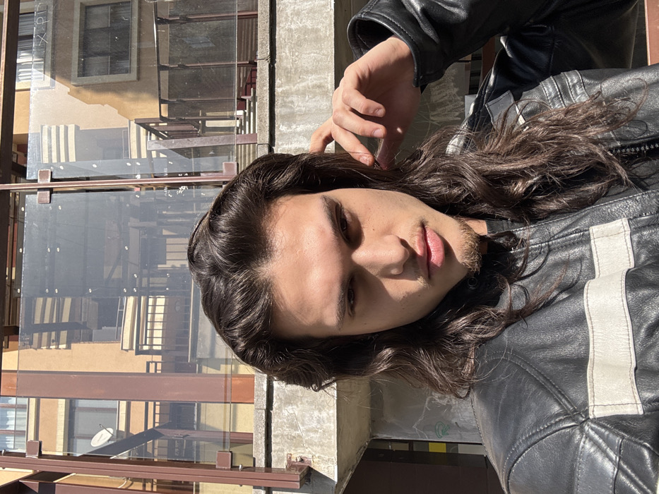 A young adult male, presumed to be in Sofia, Bulgaria, is centered in a brightly lit outdoor close-up shot. He possesses long, dark, wavy hair extending past his shoulders and exhibits light stubble on his chin and upper lip. His dark eyes are directed forward, maintaining a neutral expression. He wears a black leather jacket distinguished by two horizontal white stripes across the upper chest. His right hand is raised, with fingers lightly touching his hair near his right ear. The immediate background behind him consists of a grey concrete barrier. Beyond this barrier, a structure with dark-colored metal framing and translucent glass panels is visible, reflecting surrounding architectural elements. In the deeper background, light brown multi-story residential buildings with multiple windows, balconies, and railings are apparent, one featuring a visible satellite dish. Strong natural sunlight illuminates the scene, casting defined shadows.