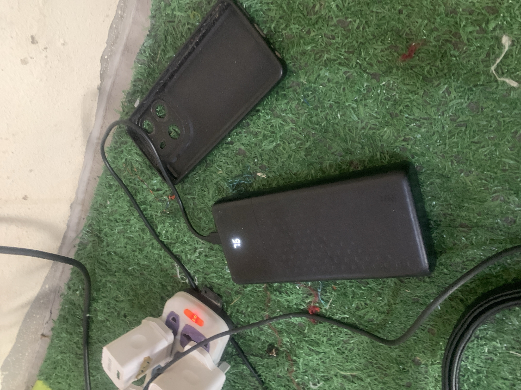In Jauro Yinu, Nigeria, on a surface covered with artificial green grass, a black power bank displays a charge of 75%. It is connected by a black cable to a white power adapter plugged into a multi-socket extension. To the right of the power bank lies a black phone case with cutouts for multiple camera lenses. The environment suggests an outdoor or indoor setting with a textured, possibly concrete or stucco wall in the background. The scene appears to be a static arrangement of electronic accessories.