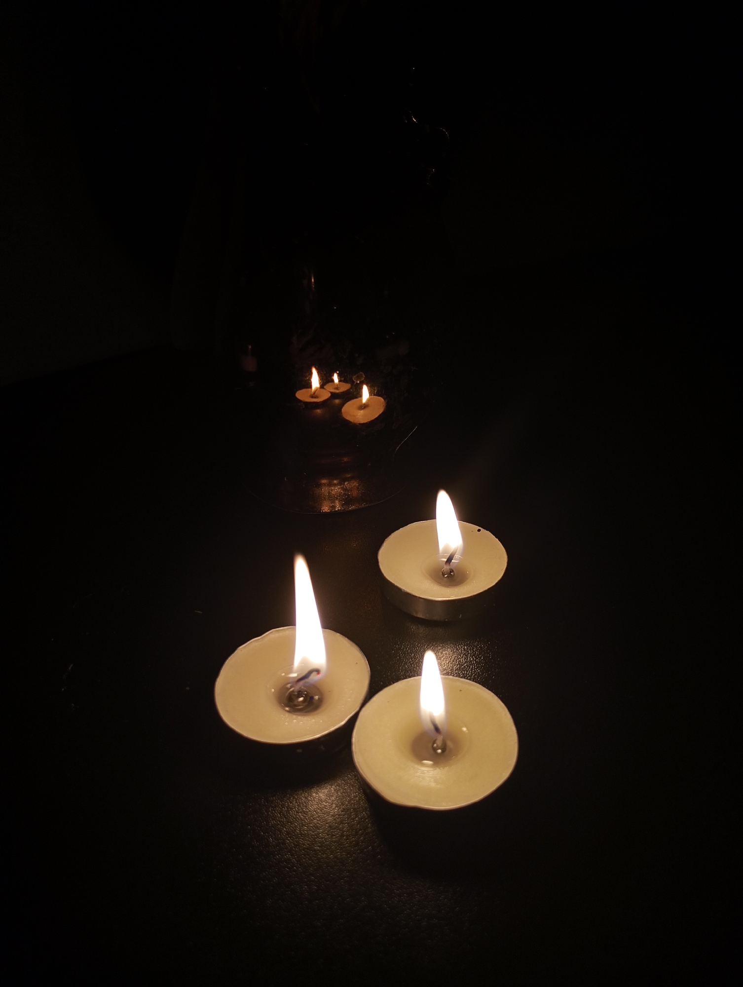 The image depicts a dark indoor setting with three lit tealight candles in the foreground. In the background, partially obscured by darkness, is a blurry object appearing to be a candle holder with multiple lit tealight candles. The setting is unknown. There are no people visible, actions or interactions shown.
