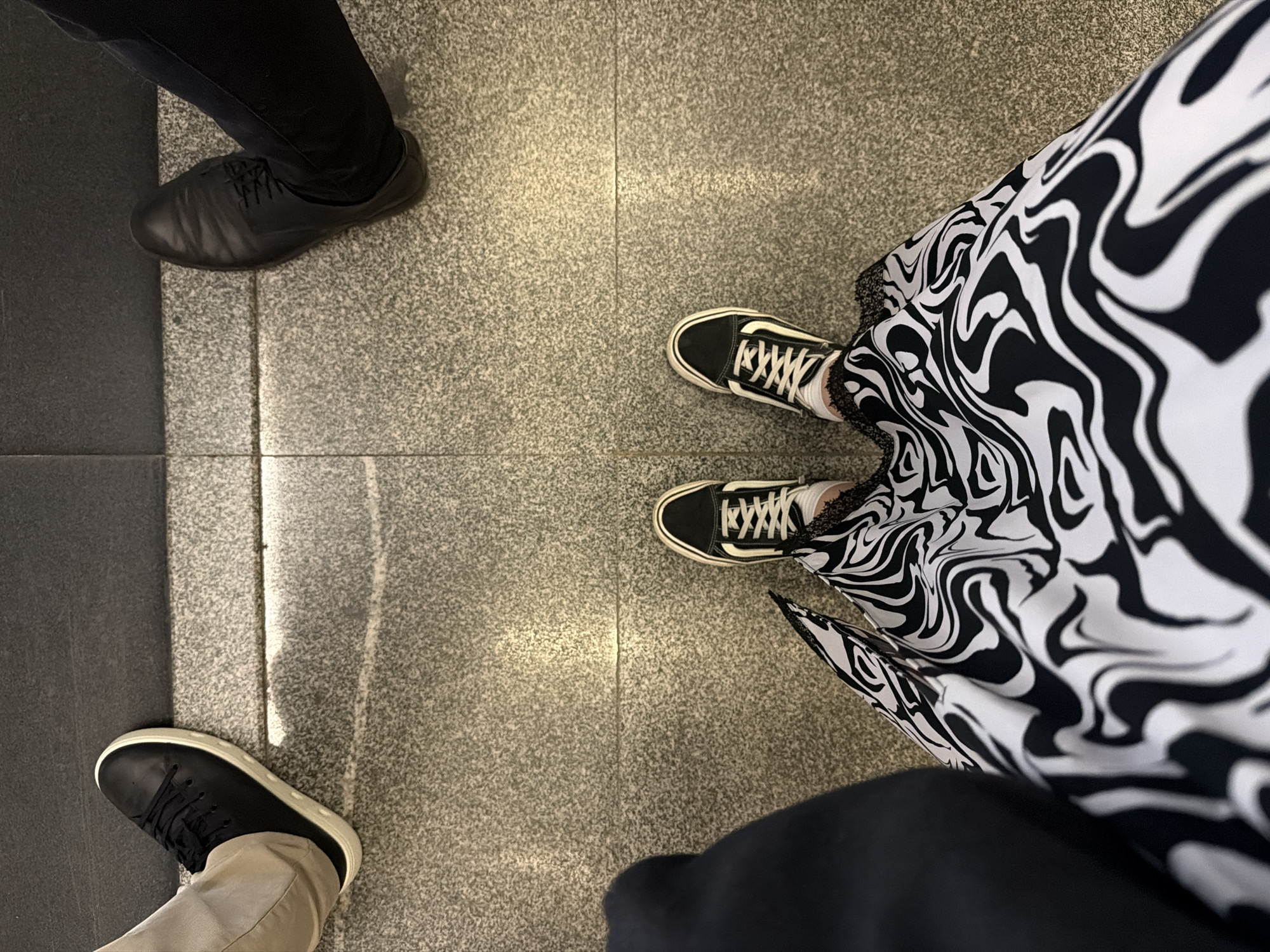 The image shows a view of the floor with three pairs of shoes and lower parts of clothing worn by three people standing on a tiled surface. The tile is light gray with a texture resembling granite. There are dark gray strips separating the tiles. The shoes on the left are black leather dress shoes and sneakers, one pair on black trousers, and one with beige trousers. On the right, the shoes are black and white canvas sneakers, and the person is wearing a black and white patterned dress. The exact location in Moscow, Russia, is not ascertainable from the image.