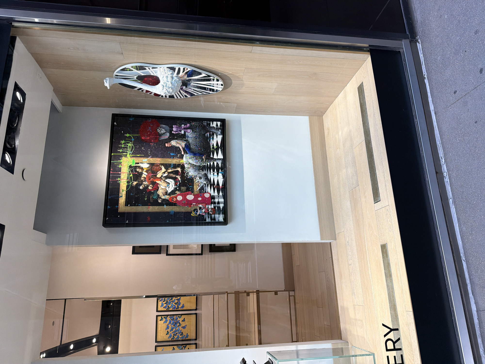 The image captures a view into a gallery in what appears to be New York City, United States. The shot is taken from the outside, looking through the gallery's window. Inside, a framed painting with a black border hangs prominently on a white wall. The painting features an elaborate scene with human figures and abstract elements, rendered in a vibrant color palette of reds, blues, and greens. A decorative object, possibly a sculpted mask or headdress in white and black, is positioned on the ceiling above the painting.
Below the painting, the gallery space is visible, with a neutral color scheme, likely beige or off-white walls. Additional framed artwork can be seen further inside the gallery, including pieces with blue butterfly designs on gold backgrounds. The floor is a light wood, possibly hardwood or laminate.
The gallery window is framed with a black border, and the exterior sidewalk can be seen to the right of the frame.