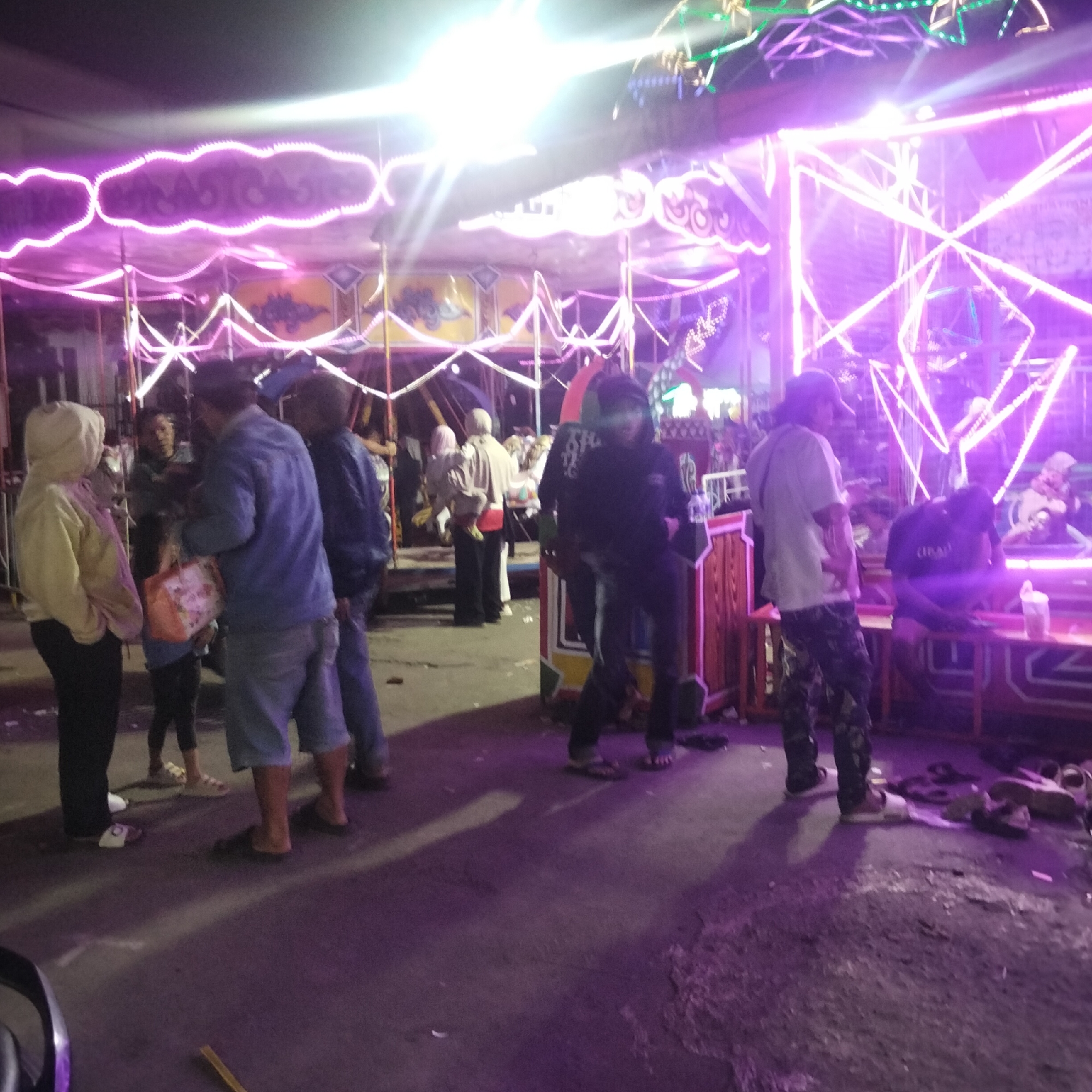 The image depicts an outdoor scene at night, illuminated by bright purple and white lights, suggesting a festive or recreational setting. In the foreground, several individuals are present. On the left, a person wearing a light-colored hooded jacket and dark pants stands facing away from the camera, holding a patterned bag. To their right, another person in a blue jacket and shorts is visible. Further right, a person wearing camouflage pants and a light-colored shirt is standing. Another individual in dark clothing is seated on a bench to the right, with a cup on the table in front of them. The background features a structure adorned with intricate neon lights, likely part of a temporary installation or amusement ride. The ground appears to be paved with some debris scattered around. The overall atmosphere is lively, with the vibrant lighting creating a sense of activity. The presence of multiple people and the illuminated structure suggest a public gathering space, possibly a fairground or market, in Cisaat, Indonesia.