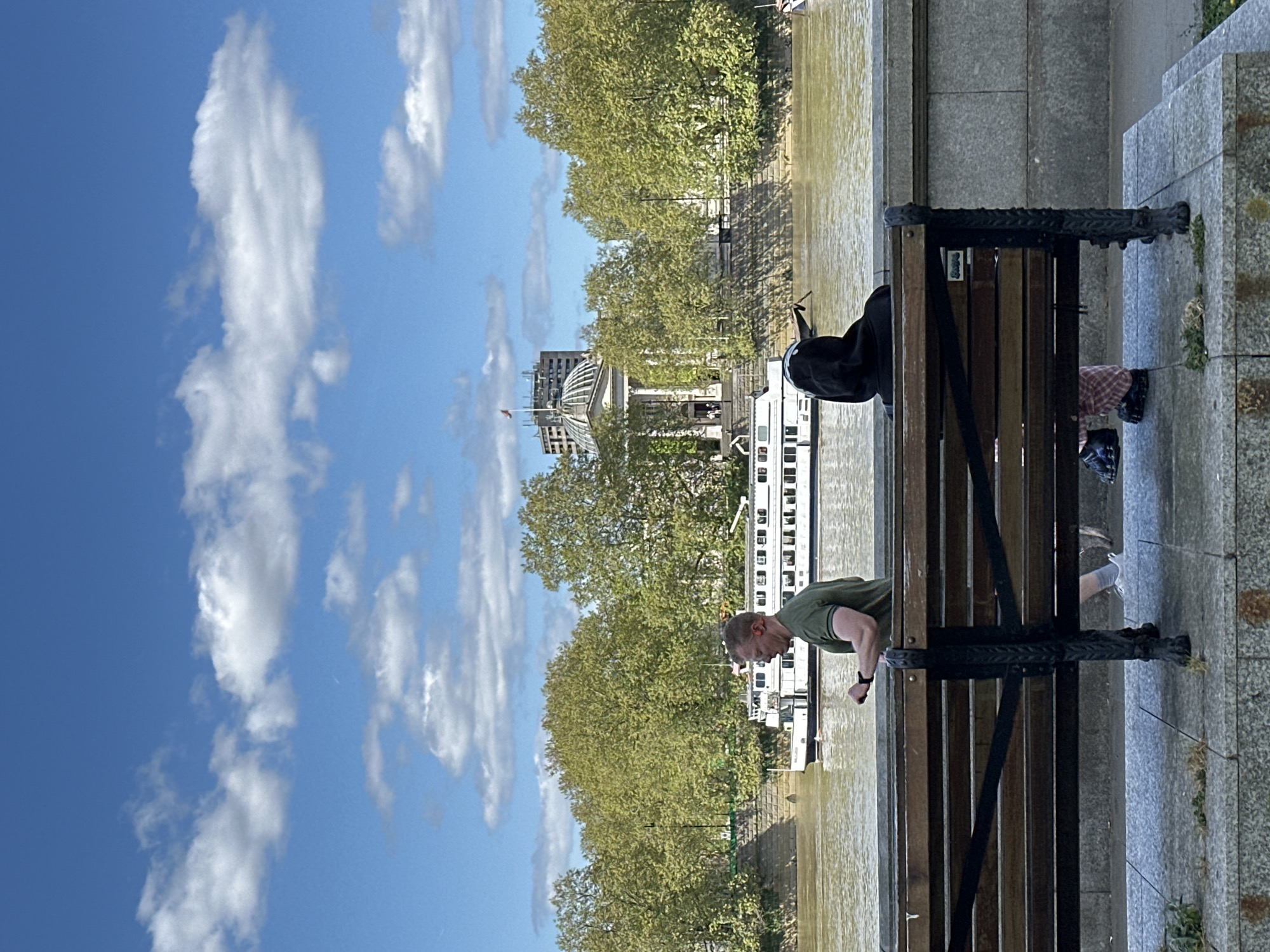 A scene in London shows a man in a green t-shirt performing push-ups on a park bench alongside the River Thames. The river is visible in the background, with a boat docked along the riverbank. A large building with a dome is behind the trees in the middle ground. The sky is blue with fluffy white clouds. The bench is made of wood and dark metal, with someone's feet and luggage on the other end. A concrete walkway with weeds is beside the bench.