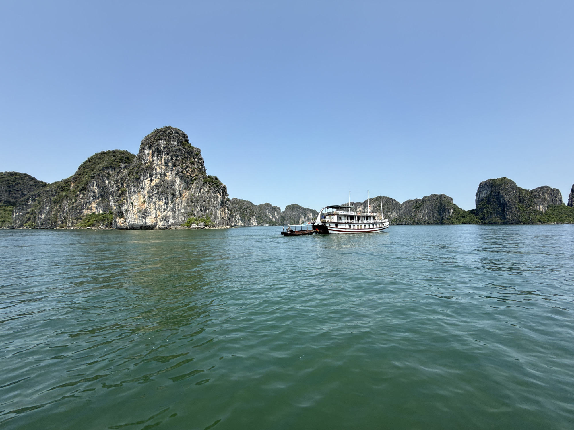 The image captures a serene daytime scene in Ha Long Bay, Vietnam, showcasing its iconic natural beauty. A wide expanse of calm, deep greenish-blue water dominates the foreground and midground, reflecting the clear, bright blue sky above. Numerous large, rugged limestone karst islands, varying in size and covered with lush green vegetation, rise dramatically from the water, forming a picturesque backdrop. The islands closest to the viewer on the left show intricate rock formations and textures.
In the center of the frame, a multi-deck white and dark-colored tourist cruise boat is either slowly moving or anchored. It features multiple windows, railings, and covered upper decks, indicating facilities for passengers. A smaller, darker utility boat or tender is positioned behind and to the left of the main cruise boat, seemingly attached or in close proximity. No people are visibly depicted in the scene.
The bright sunlight suggests it is midday or early afternoon, with perfectly clear weather conditions. The overall atmosphere is tranquil and scenic, typical of a tourist excursion in this famous Vietnamese location. On the side of the larger boat, the text "HP. SEA STAR LEGEND CRUISE" is visible.