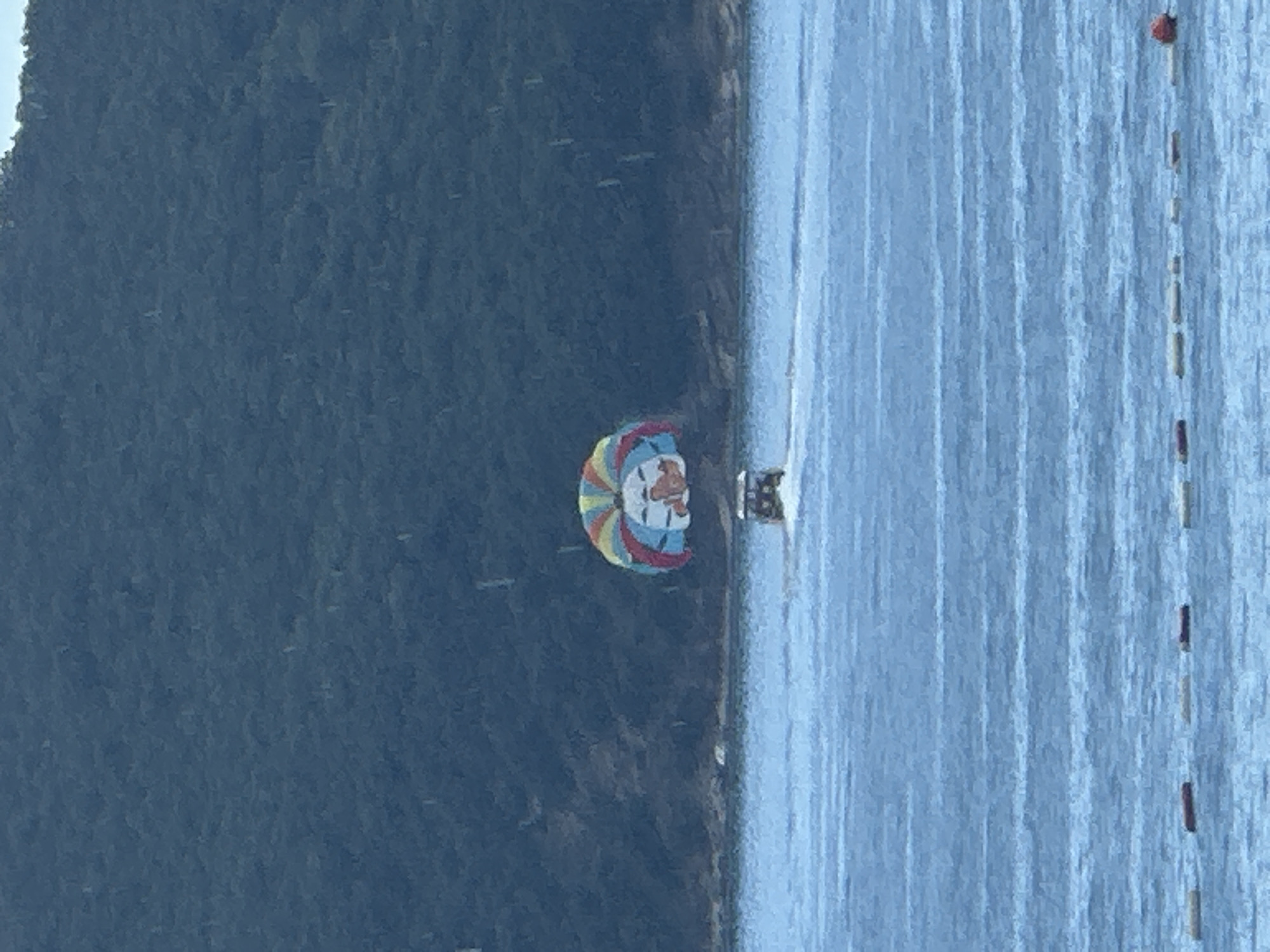 The image shows a parasailer in the water off the coast of Rafailovići, Montenegro. The parasailer is a colorful umbrella-shaped parachute with a depiction of a person's face. They appear to be attached to a boat, though the boat is not fully visible. The sea is calm with light blue and white rippling water. The background is dominated by a dark, forested hill on the left side of the image, suggesting a mountainous coastline. Along the right edge of the water, a series of buoys are visible, marking a boundary or a course. The overall scene suggests a recreational activity on a clear day, likely during daylight hours. The image is oriented vertically, with the hill on the left and the water on the right. No visible text is present in the image.