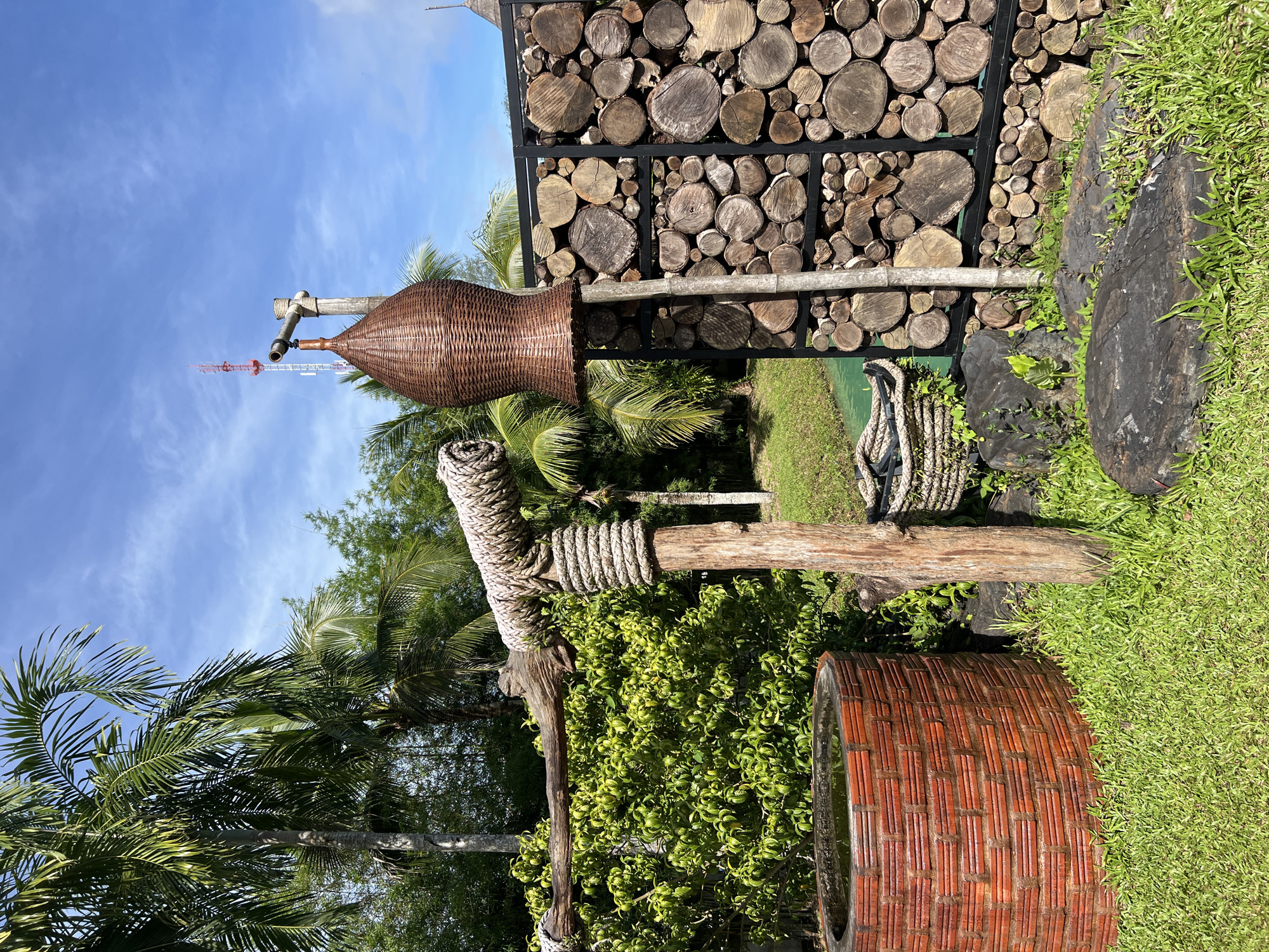 The media file displays an outdoor scene in a tropical setting, likely Thailand, given the presence of lush palm trees and dense green foliage. The image is oriented vertically, giving a sense of looking up.
In the foreground, there are several rustic, naturalistic decorative elements. A large, woven basket, shaped like a wide-mouthed bottle, is supported by wooden posts wrapped with thick rope. Another similar rope-wrapped wooden post is visible below it. To the right, a large, round, terracotta-colored planter with a woven pattern sits on vibrant green grass. The planter appears to be empty or filled with something reflective that mirrors the sky.
In the background, a wall constructed from stacked, circular wooden logs of varying sizes is a prominent feature. This wall is framed by dark metal supports. Beyond the wall, more tropical vegetation and palm fronds are visible, suggesting a garden or natural park setting. A tall, red and white lattice tower, possibly a communication tower, is visible in the distance against a bright blue sky with scattered white clouds.
The time of day appears to be daytime, with clear blue skies and bright sunlight casting shadows, indicating fair weather. There is no text visible in the image. The overall impression is one of a serene, natural, and possibly tropical garden or outdoor recreational area.
