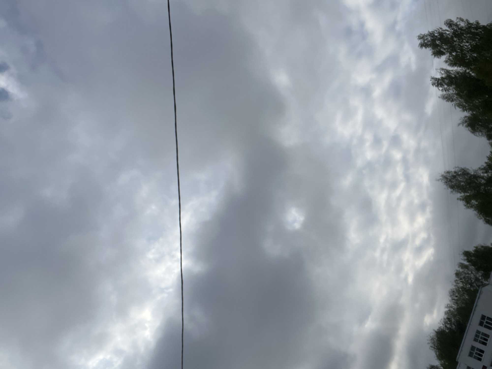 The image shows a cloudy sky with a power line running vertically. On the right side, there are trees and the corner of a building with windows. The location is likely in Chaikovsky, Russia, though there are no identifying features to confirm the location. There are no people visible.