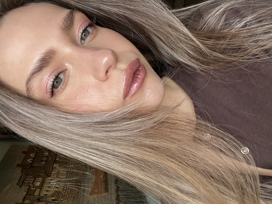 A female subject is positioned in a close-up, chest-up portrait shot, looking directly into the camera. She has long, blonde hair with lighter highlights, parted on the side and draped over her shoulders. Her eyes are light-colored (appears blue/green), and her eyebrows are defined. She is wearing pink eyeshadow with a subtle winged eyeliner and glossy pinkish-brown lipstick. Small, circular silver stud earrings are visible on her earlobes, partially obscured by her hair on the left side of the frame. She wears a dark brown long-sleeved top with a round neckline. In the background, to the left of the subject, a wall tapestry or artwork is visible, depicting an old-world cityscape with buildings featuring pitched roofs and dormer windows, alongside a boat on a body of water. The setting is an indoor location in Saint Petersburg, Russia.