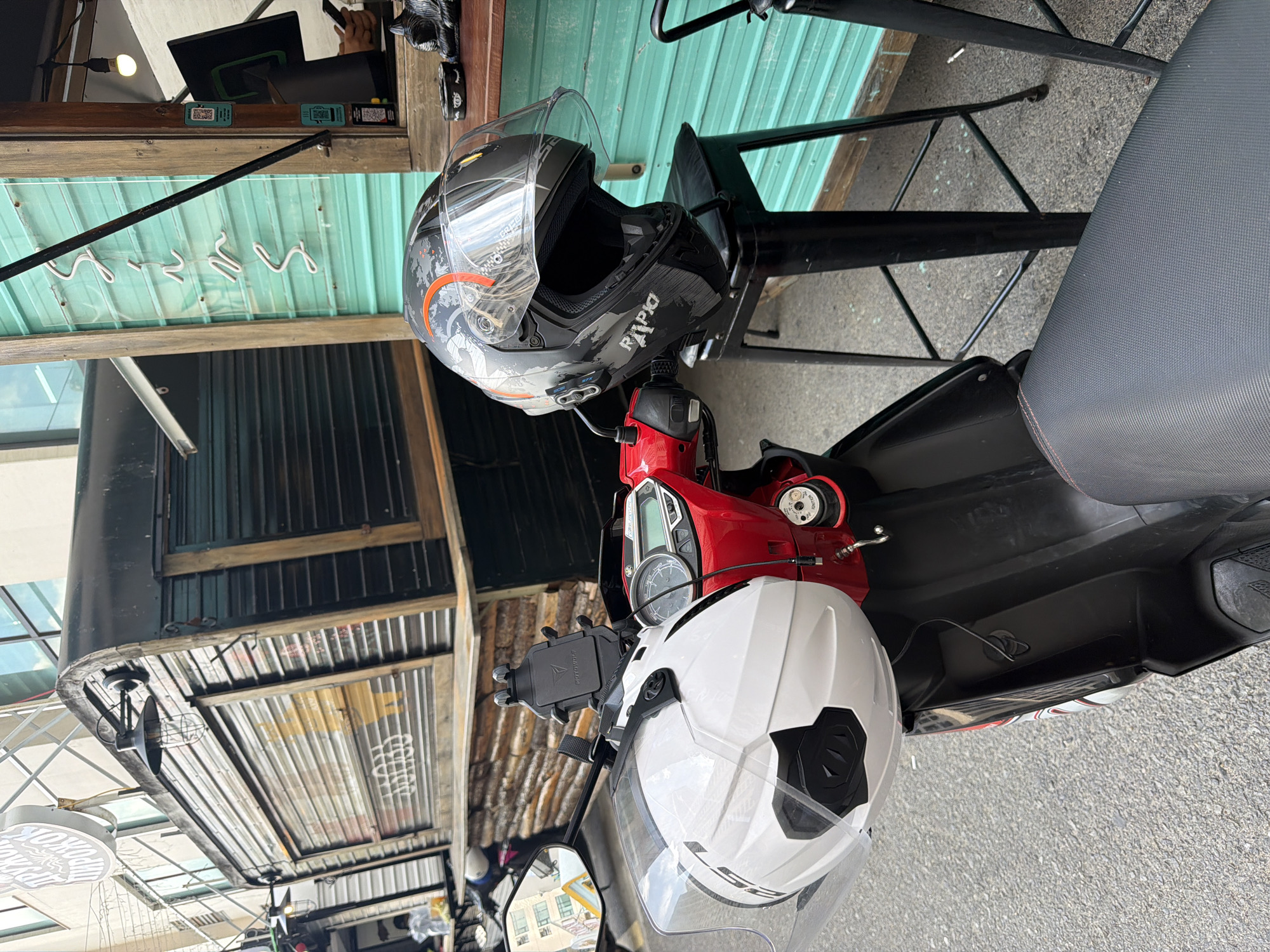 The image shows a parked red scooter with two helmets resting on it, taken from an elevated perspective. The upper helmet is black with orange and grey detailing, while the lower helmet is white with black accents. The scooter is parked on a grey asphalt surface. Overhead, there is a building with a corrugated metal roof and a light green awning. The location is suggested to be Bac Nha Trang Ward, Vietnam. There are no people visible in the immediate frame.