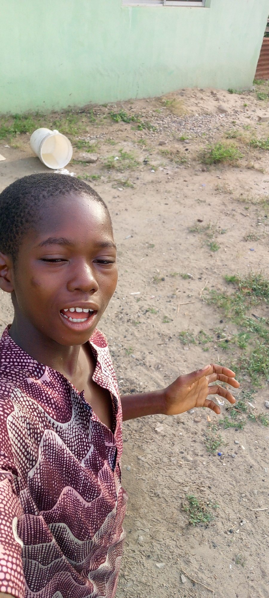 A person, likely a young male, is standing in an outdoor area. He has dark skin, short black hair, and is wearing a patterned button-up shirt. He is speaking or smiling, with his mouth open, and his right hand is partially visible. The environment appears to be a dirt or gravel area with some sparse vegetation. In the background, there is a building with light green walls and a window. A white bucket lies on its side on the ground. The location is estimated to be in Adankolo, Nigeria.