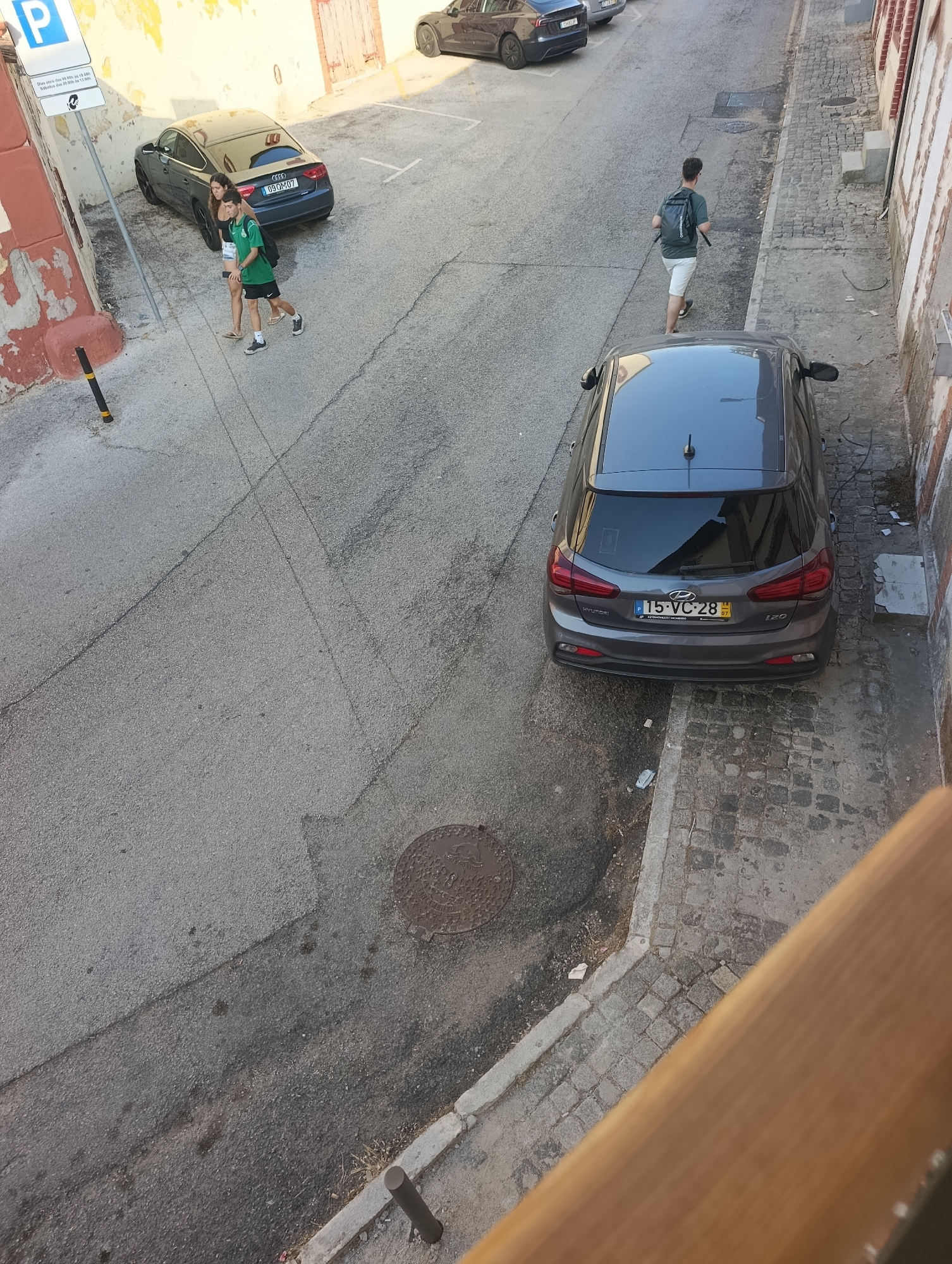 The image depicts a street scene in Setúbal, Portugal. In the foreground, a wooden railing is visible, out of focus. The middle ground shows a grey Hyundai i20 parked on the right side of a paved street. The car has the license plate "15-VC-28". A person wearing a dark green t-shirt and white shorts, carrying a backpack, walks away from the car down the street. Further up the street, a dark grey Audi is parked on the left side. Two individuals, a woman wearing a black top and denim shorts, and a man in a green t-shirt and black shorts, are walking towards the Audi. The street is lined with buildings on the right and a wall with peeling paint on the left. The ground is paved with asphalt and cobblestones in some areas. A manhole cover is visible on the street. The lighting suggests it is daytime, with shadows present.