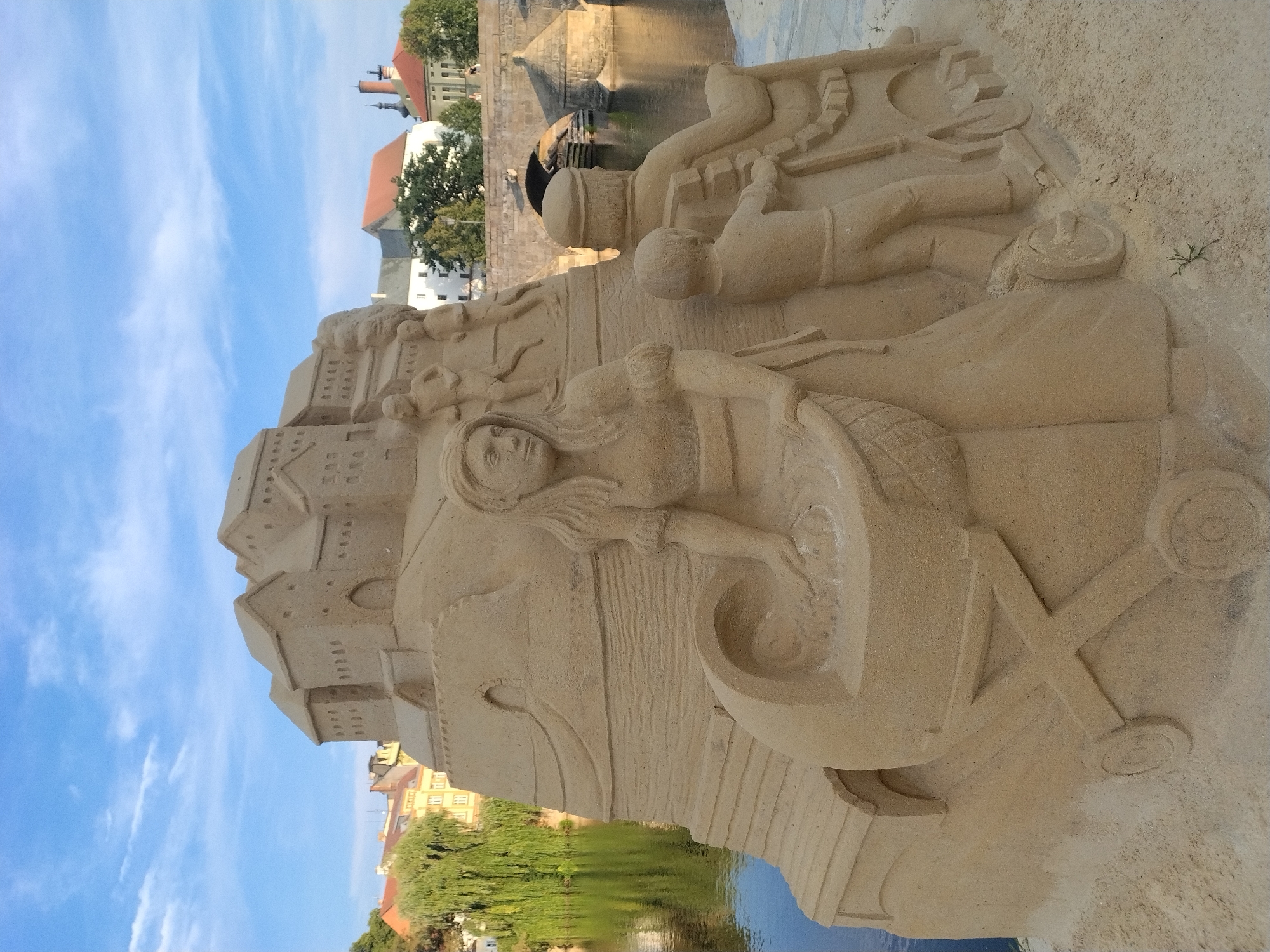 The image depicts a sand sculpture on the bank of the Otava River in Písek, Czechia. The sculpture features a central motif of a family, including a woman holding a baby in a cradle, a man, and a child. Behind them, a sandcastle structure is sculpted, evoking a historical or fantastical setting. In the background, a stone bridge with arches spans the river, and buildings of Písek are visible on the opposite bank, including trees and a church spire. The sky is blue with scattered white clouds. The scene is brightly lit, suggesting daytime.