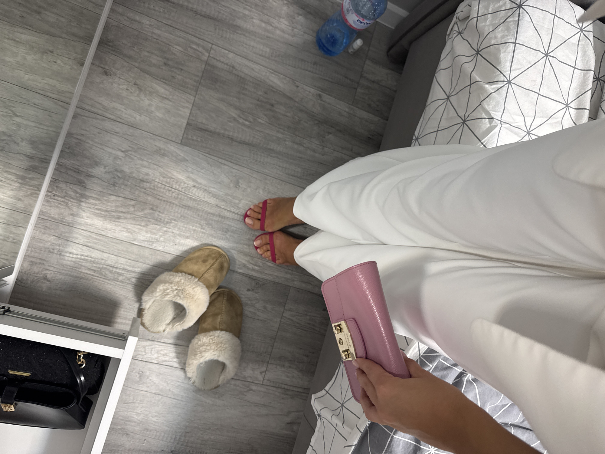 The image shows an indoor scene, possibly a bedroom or dressing area. The foreground features a person wearing white trousers and pink sandals, holding a pink leather clutch. Near the person's feet are a pair of brown and beige fur-lined slippers. A water bottle is placed on the floor near the head of the bed.
The room has a gray wooden floor, a white bedside table with a black purse or bag on the shelf, and a bed with a gray and white patterned duvet.
The location cannot be determined definitively from the image alone. No explicit details refer to Burgas, Bulgaria.