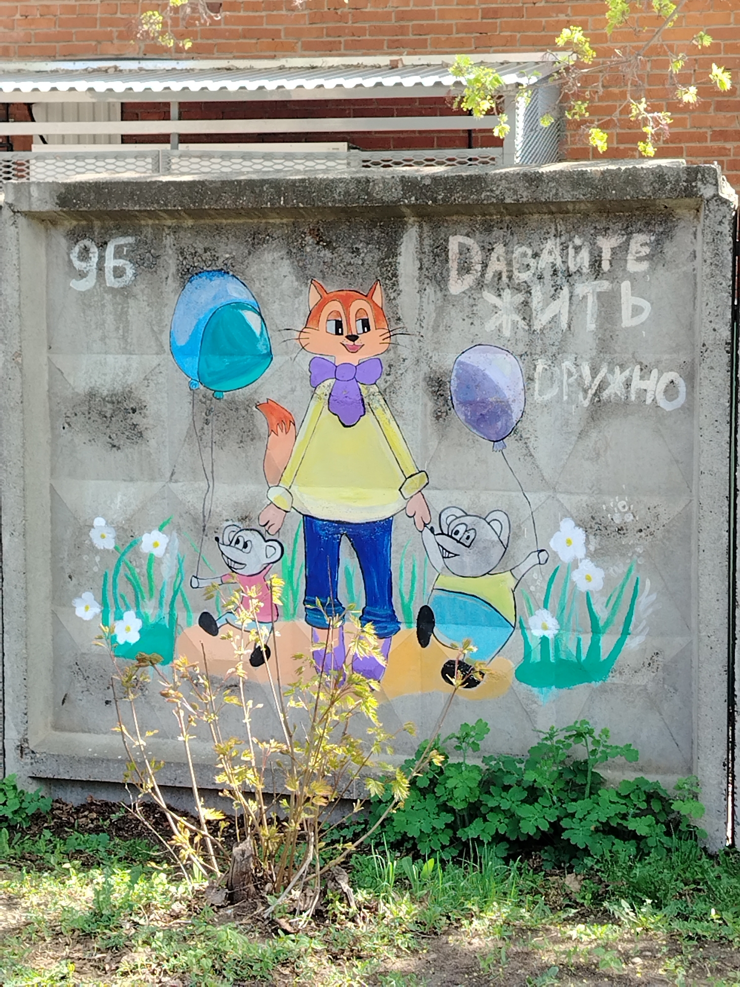 The image depicts a concrete fence, likely in a residential area, featuring a mural-style painting. The fence is painted gray and appears to be a precast concrete panel. The mural depicts a stylized orange fox wearing a yellow shirt, blue pants, and a purple scarf, standing between two mice. Each mouse holds a balloon; one is blue, and the other is purple. Green grass and white flowers are painted around the feet of the cartoon characters. To the upper left of the fox is the number "96" painted in white, and to the upper right is text in Russian: "ДАВАЙТЕ ЖИТЬ ДРУЖНО" (Let's Live Together). In front of the fence, at ground level, are patches of grass and other greenery. Part of a brick building and a metal railing structure are visible in the background. The image is taken during daylight hours in Noviy, Russia.