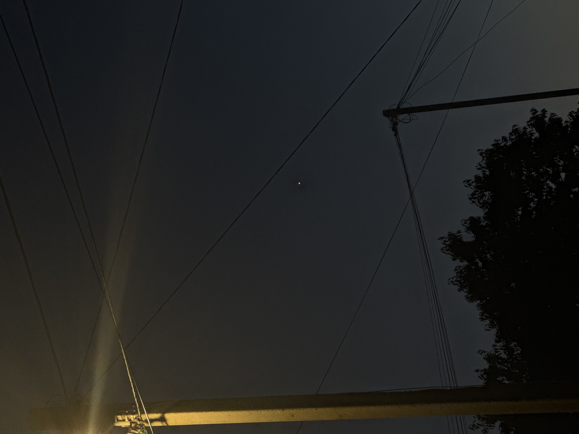 The image depicts a nighttime scene in Mikhaylovsk, Russia. A concrete utility pole, illuminated by a yellowish light from the top, stands on the left side of the frame. Several overhead power lines crisscross the dark sky. In the upper middle of the sky, a single, bright star is visible. Below the power lines and in the lower part of the image, a silhouette of trees forms a dark mass against the dim sky. Another utility pole, silhouetted and with tangled wires, is positioned in the lower right quadrant. The overall atmosphere is one of a quiet, ordinary evening in a residential or semi-rural area.