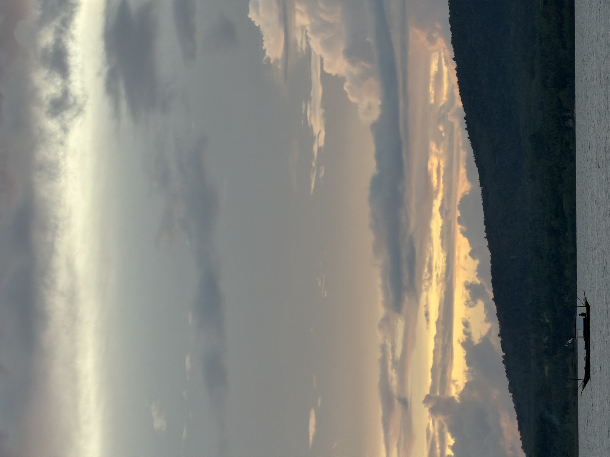 A lone, dark-hulled boat with a single person visible aboard sails on calm, dark water. The boat has a raised stern and a small flag. It is positioned in the lower-left quadrant of the frame. Behind the water, a silhouetted, tree-covered hillside stretches horizontally across the image. The sky is filled with dramatic clouds, with a bright, yellowish-orange band of light near the horizon, suggesting either sunrise or sunset. The sky transitions to darker grays and blues higher up, with lighter, wispy clouds interspersed. The overall impression is of a serene, natural landscape at twilight. The image appears to be a photograph of the sea and coast of Na Thung, Thailand.