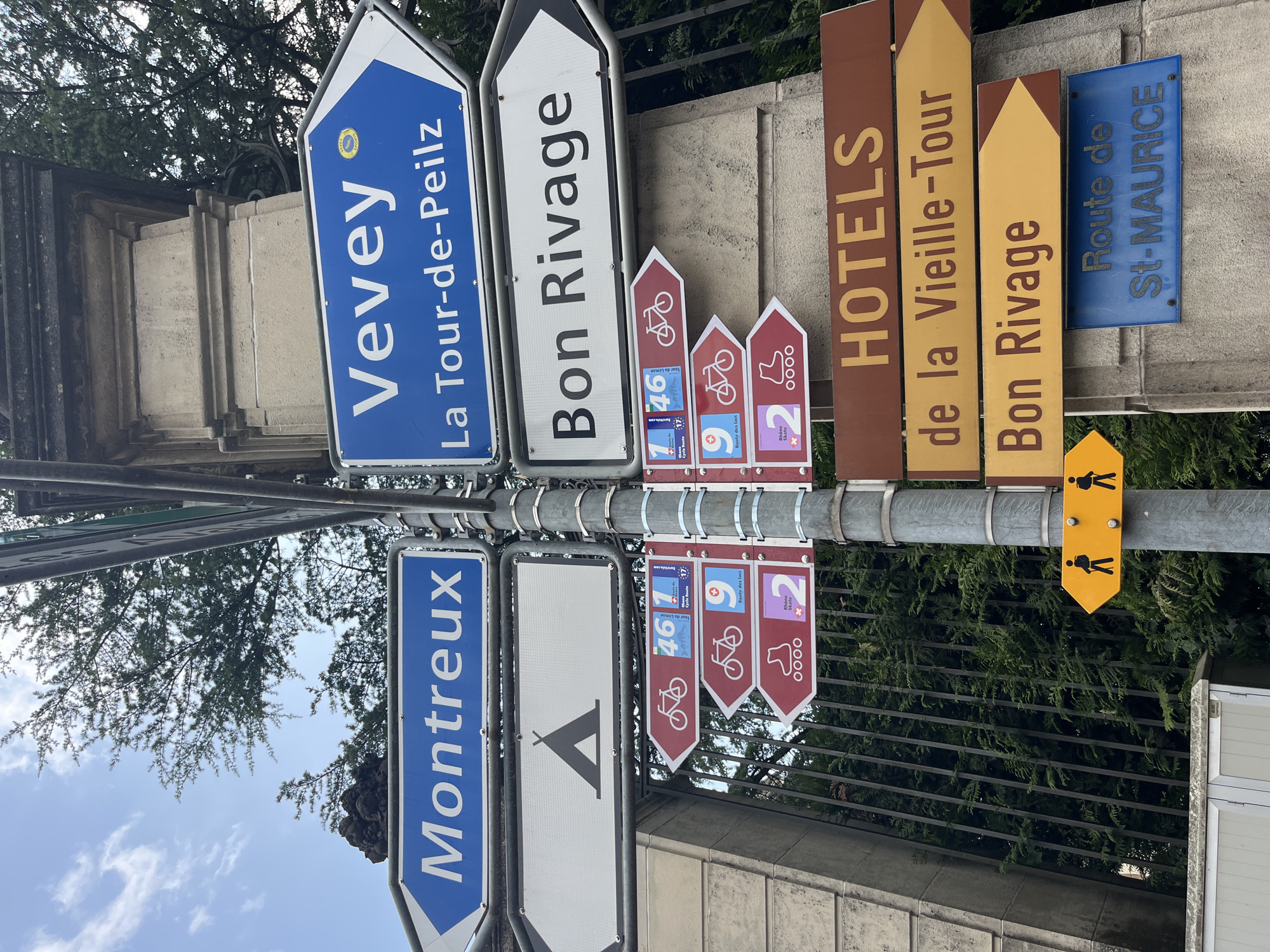 The image shows a directional signpost in Montreux, Switzerland, with multiple signs pointing in various directions. The signs indicate destinations such as "Montreux", "Vevey", "La Tour-de-Peilz", "Bon Rivage", and "St-Maurice". There are also signs with symbols for camping, cycling (routes 46, 1, and 9), and rollerblading (route 2). A sign for "HOTELS" and "de la Vieille-Tour" is also visible. A yellow directional sign with two pedestrian figures is present. The signpost is mounted on a pole next to a stone pillar and a metal fence, with trees and a cloudy sky in the background.