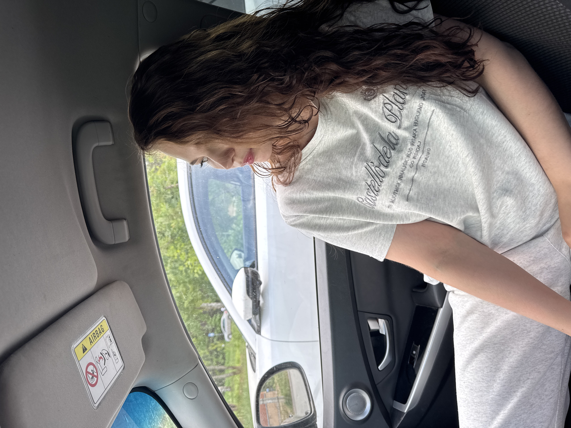 A young person with long, curly brown hair is sitting in the passenger seat of a car. The person is wearing a light gray t-shirt with text and the phrase "Castelli dela Plan" on it, and light gray sweatpants. The car is parked, as another white vehicle is visible through the passenger window. The car's interior includes a gray sun visor with an airbag warning label and a gray door panel. The location in the city of Chaikovsky, Russia, is not discernible from the image.