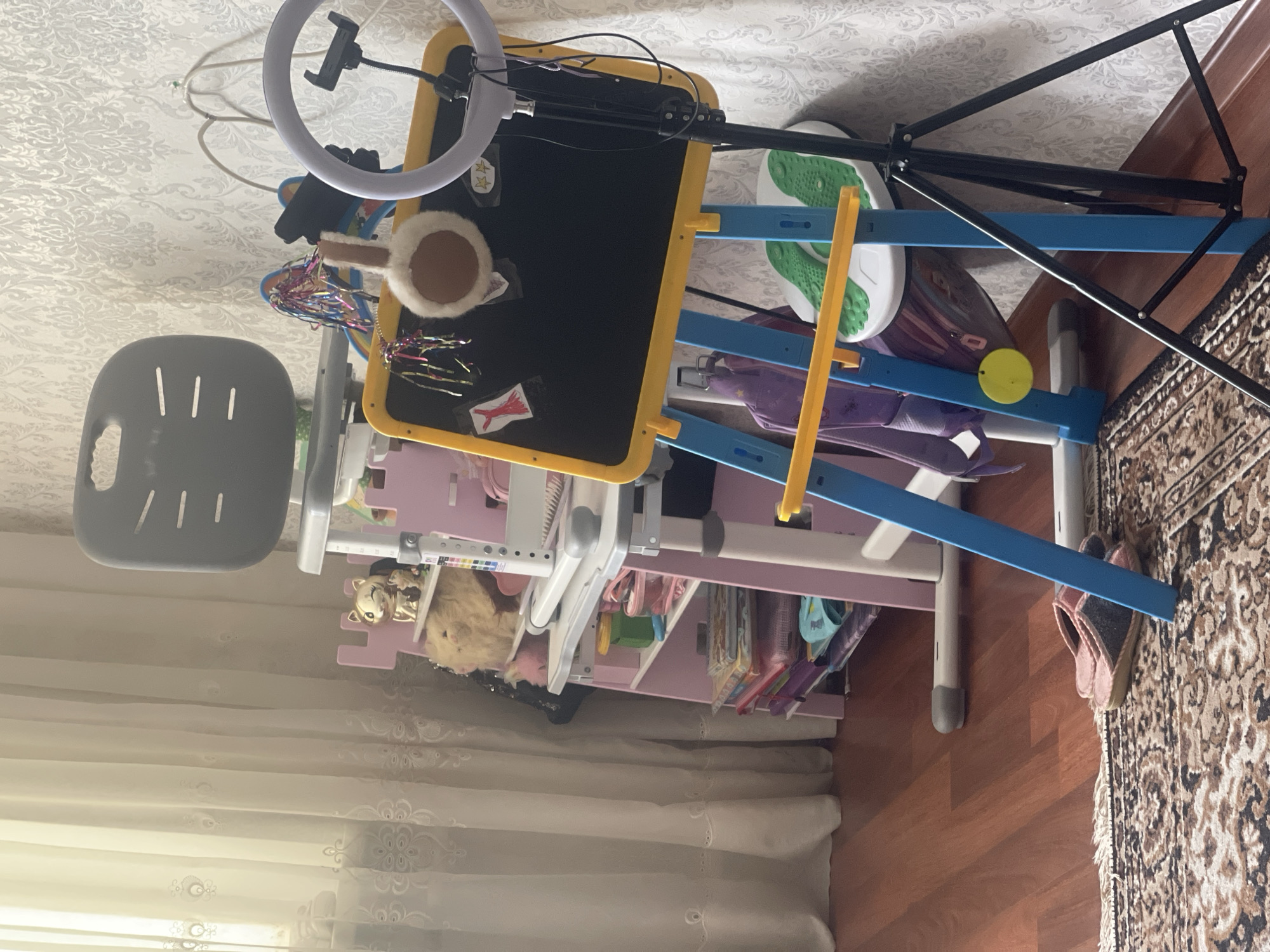 The image displays an indoor scene, likely a child's room or activity area, in the city of Unknown, Unknown. To the left, a window is covered by sheer white curtains featuring embroidered patterns, admitting natural light. The background wall is covered with a light-colored, ornate patterned wallpaper. In the central midground, a children's adjustable desk and chair set is present. The chair is grey with a slotted backrest. The desk features a pink frame with white surfaces and integrated side shelves. These shelves contain a golden winking cat figurine, several plush toys, a stack of books, and small storage containers, including a green one and a pink one.
To the right of the desk, a children's easel with a blackboard surface, framed in yellow and supported by blue legs, is positioned. Items affixed to the blackboard include a red paper dress, a white fluffy circular object, and two star shapes. A purple backpack with a yellow circular tag is suspended from one of the easel's blue legs. Adjacent to the easel, a black tripod stand supports a white ring light equipped with an adjustable phone holder.
The floor is polished hardwood, with a section of a brown and beige patterned rug visible in the foreground on the left. A pair of pink and grey slippers rests on the rug. Behind the easel, a white and green foot massager or exercise device is visible on the hardwood floor. No individuals are present in the scene.