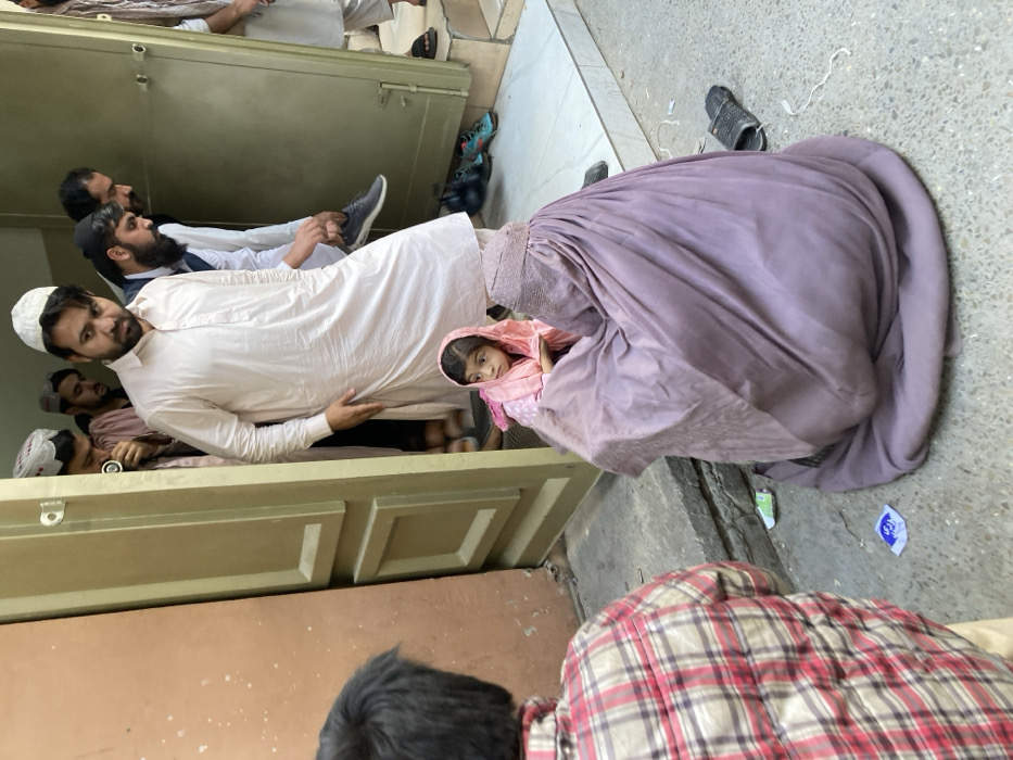 In Quetta City Tehsil, Pakistan, an outdoor scene unfolds at the entrance of a building. A young boy in a red and white plaid shirt stands with his back to the viewer in the lower left. To his right, a woman clad in a light purple full burqa (chadri) is seated on the grey concrete ground, her back partially towards the viewer. Adjacent to her, a small girl, approximately 3-5 years old, wearing a pink headscarf and clothing, also sits on the ground, looking towards the left of the frame. Directly behind the girl and woman, a man in a light-colored shalwar kameez and a white cap stands within a doorway, looking intently towards the left. Behind him, several other adult males are visible inside the building's entrance. One man is observed holding a pair of dark sneakers, while another wears a traditional cap. Scattered footwear, including dark slippers and sneakers, is present on the concrete ground near the doorway's raised threshold. The doorway consists of a brown wooden frame on the left and a lighter green wall section on the right. Small pieces of debris, including wrappers and white string-like material, are visible on the concrete ground. The area is lit by natural daylight.