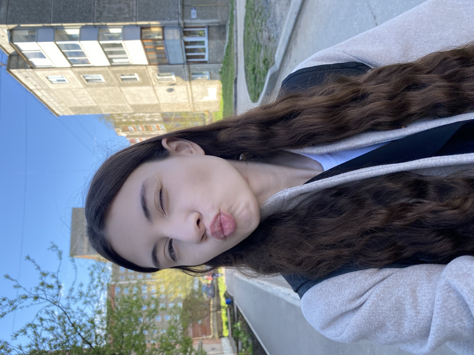 A young woman is photographed outdoors, likely in Votkinsk, Russia, based on the architecture and background. She is oriented at an angle to the camera, with a pouting expression and one eye partially closed in a winking gesture. She has dark hair and fair skin, and is wearing a light-colored hoodie with dark trim. The background shows a multi-story concrete apartment building, common in Russian urban areas, along with greenery, including trees and grass. The image appears to be taken during daylight hours under natural lighting conditions. A concrete walkway or ledge is also visible.