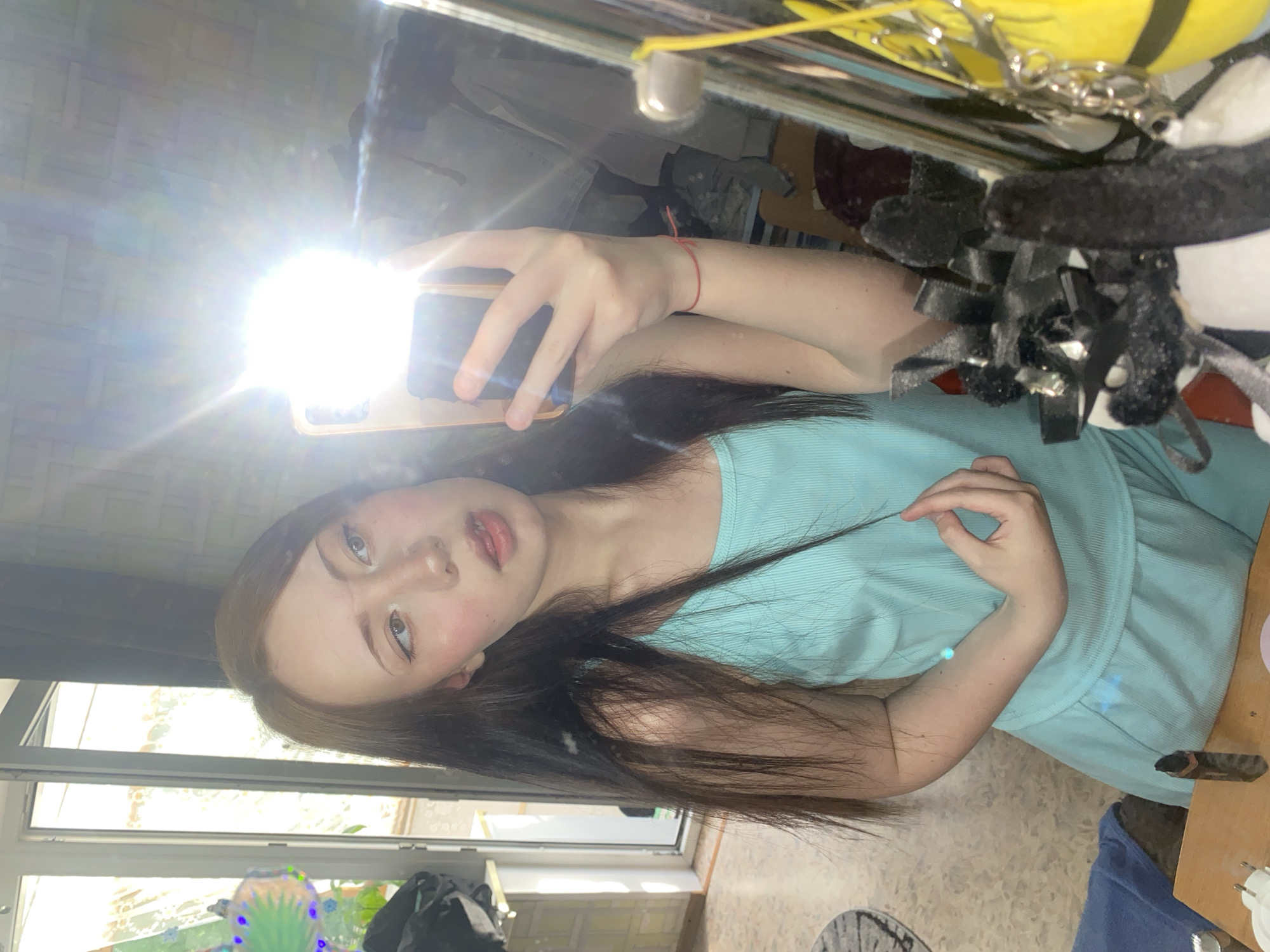 A young person with long, dark hair is taking a selfie in what appears to be an indoor setting. They are wearing a light blue top. The location is unclear but seems to be a home environment, possibly near a window. A bright light, likely from the phone's flash, obscures some of the details. Other objects are visible in the background, including a mirror and what appears to be personal belongings.