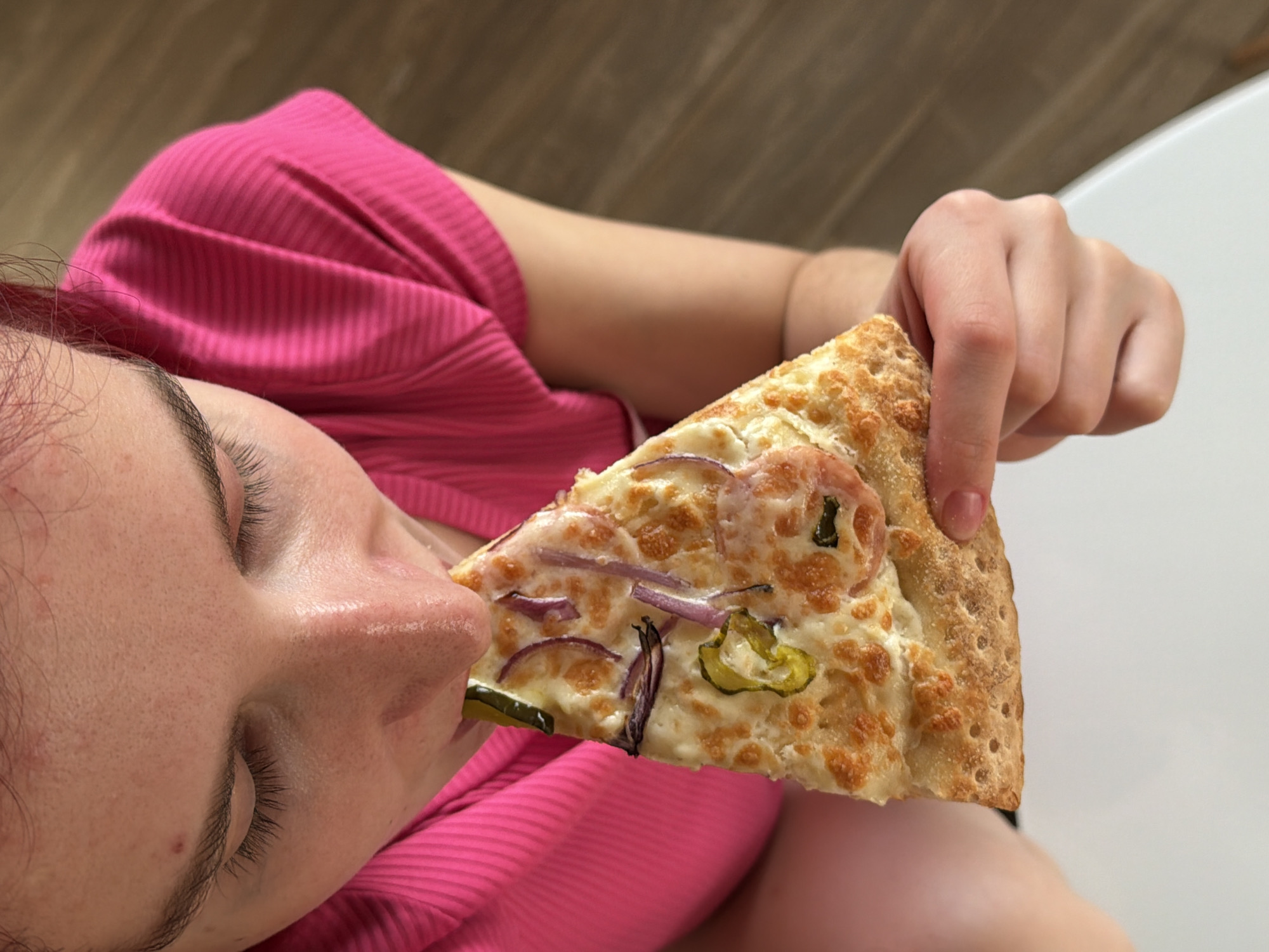 A woman wearing a pink top is about to eat a slice of pizza. The pizza has cheese, red onions, and sliced green peppers on it. The background is blurry, but there appears to be a white bathtub in the distance. There is no definitive evidence to support that the image was taken in Chaikovsky, Russia.