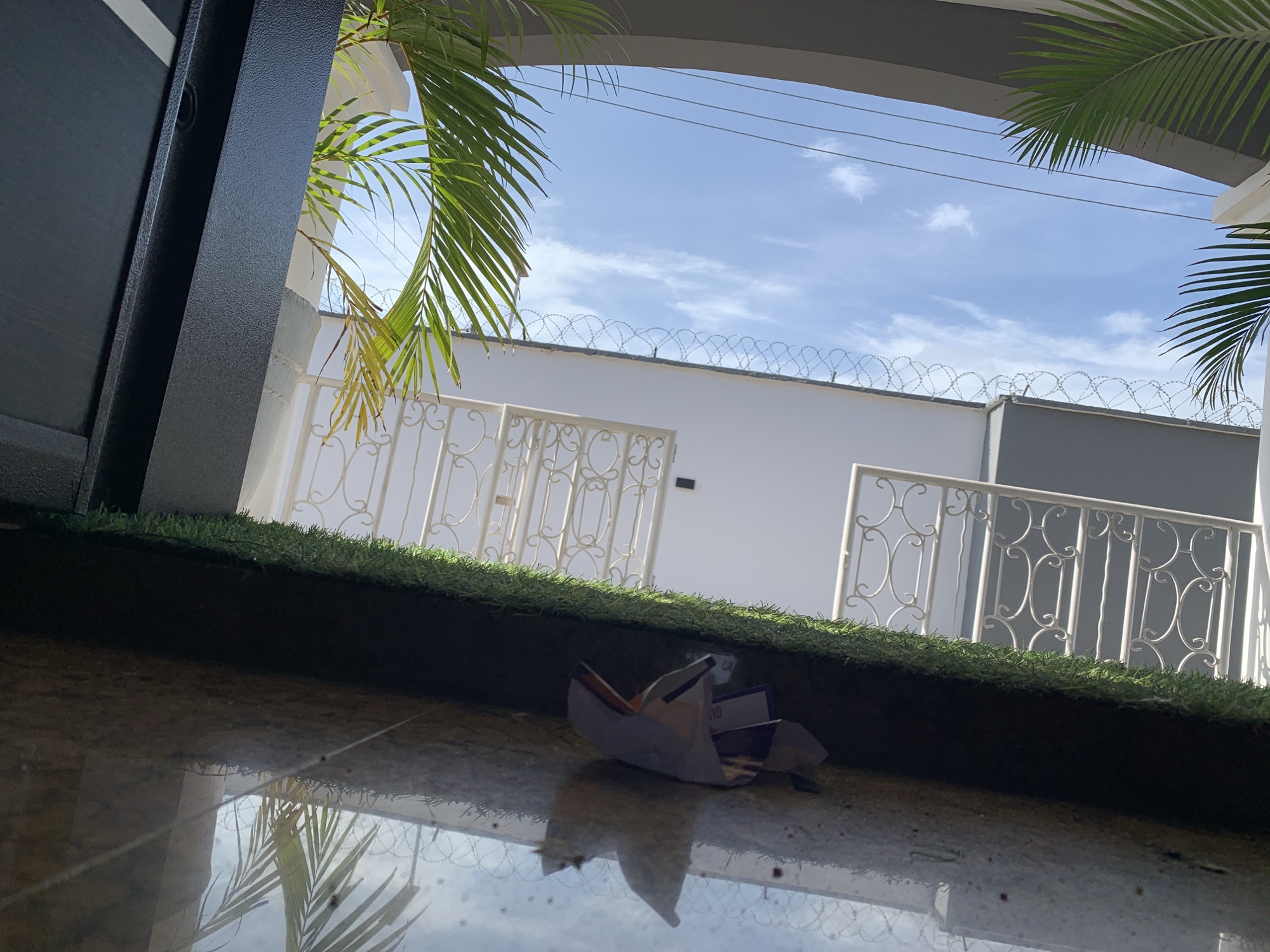 The image shows a view of the sky and buildings from a low angle in Gura Top, Nigeria. In the foreground, a wet, reflective surface, possibly a tiled floor or a puddle, shows reflections of palm fronds and some debris. A small, partially unfolded origami boat made of colorful paper lies on this wet surface. Above this, a strip of artificial turf is visible. In the middle ground, there are white decorative metal railings and a light-colored wall. Above the wall, barbed wire is strung along its top. In the background, a bright blue sky with scattered white clouds is visible, along with power lines. Lush green palm fronds extend into the frame from the left and right sides, partially obscuring the view of the sky and buildings. The overall scene suggests an outdoor setting, likely a residential or commercial area, possibly after rain given the wet surfaces.