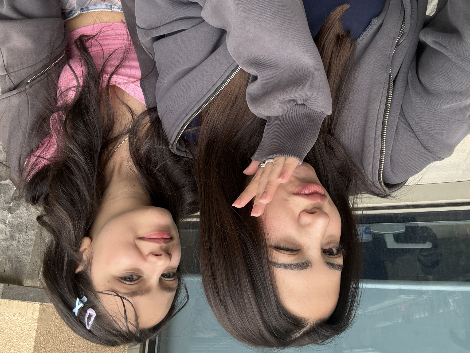 Two young women are pictured in an upside-down orientation. The woman on the left has brown hair, light skin, and is wearing a pink top under a gray zip-up sweatshirt. She has hair clips in her hair. The woman on the right has brown hair, light skin, and is wearing a gray zip-up sweatshirt. She has a ring on her finger and is winking. The background includes a window, a curb, and a building. The location is likely in a city, possibly Yerevan, Armenia.