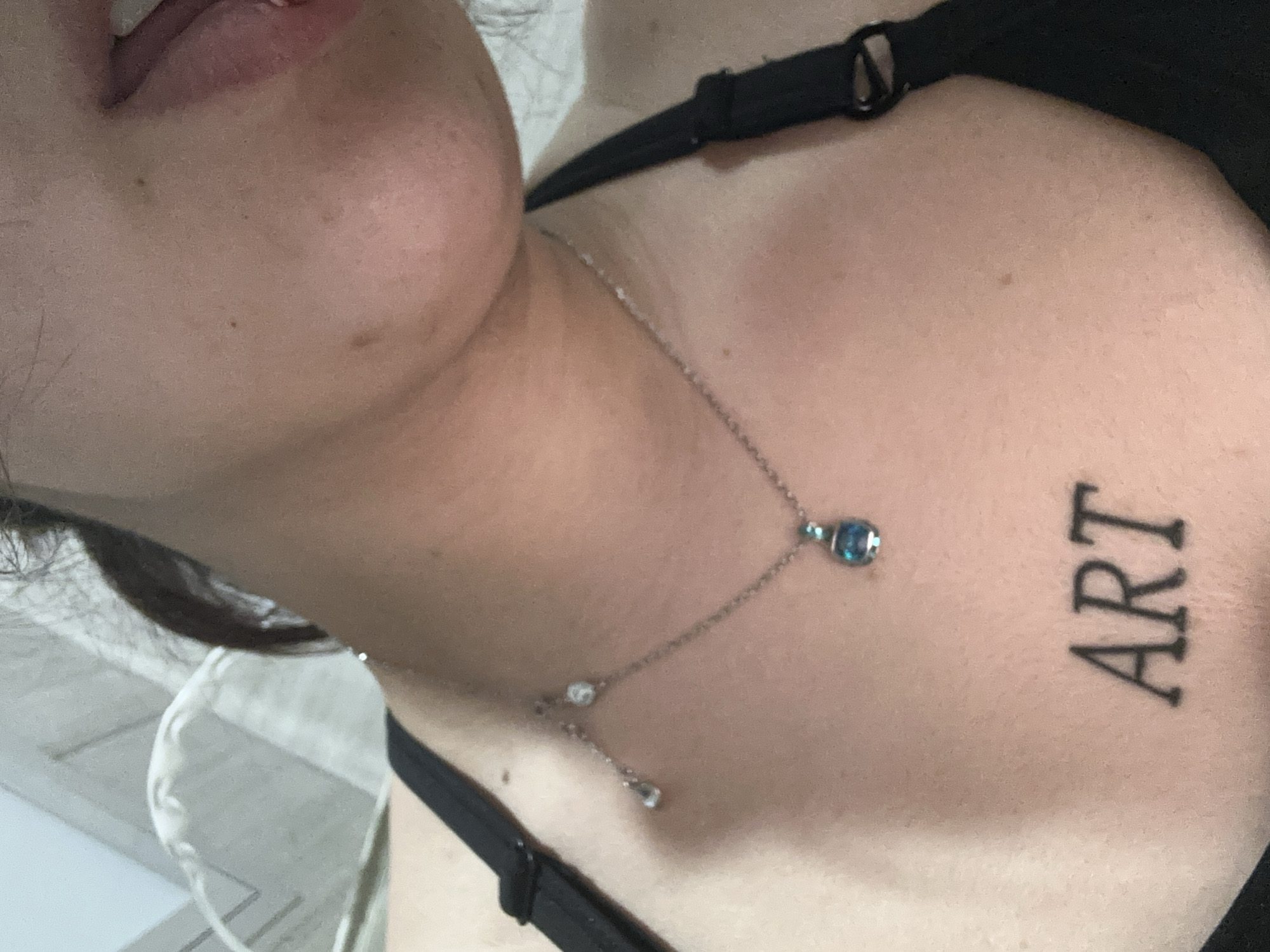 A close-up, angled shot of a person's upper chest and face. The person has fair skin, and the angle emphasizes their jawline. They are wearing a black bra or top. A delicate silver necklace with blue and clear gemstone accents is visible. The word "ART" is tattooed in black, block letters on their upper chest. The background is indistinct, but possibly indoors. There is no indication of the city of Sofia, Bulgaria in the image.