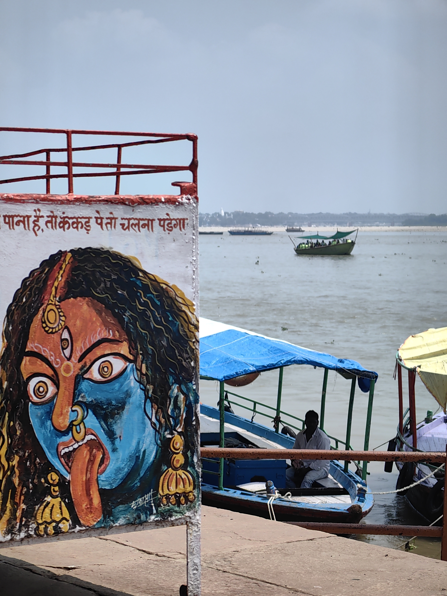 A foreground painting of the goddess Kali is on a white wall with red railings. The painting depicts Kali with blue skin, wide eyes, a third eye on her forehead, and a long orange tongue sticking out. She is adorned with large yellow earrings and a nose ring. Above the painting, Hindi text is visible in red lettering.
In the middle ground, a light blue boat with a blue awning is docked. A man in a white shirt and light-colored pants sits in the boat, holding something in his hands. Another boat with a yellow awning is partially visible to the right. The boats are on the Ganges River, a prominent river in Varanasi, India.
In the background, several other boats are visible on the water, and a cityscape with buildings and bridges stretches along the distant shore. The sky is overcast with a light blue hue. The overall scene suggests a location along the ghats of Varanasi, a popular destination for boat rides and religious activities.