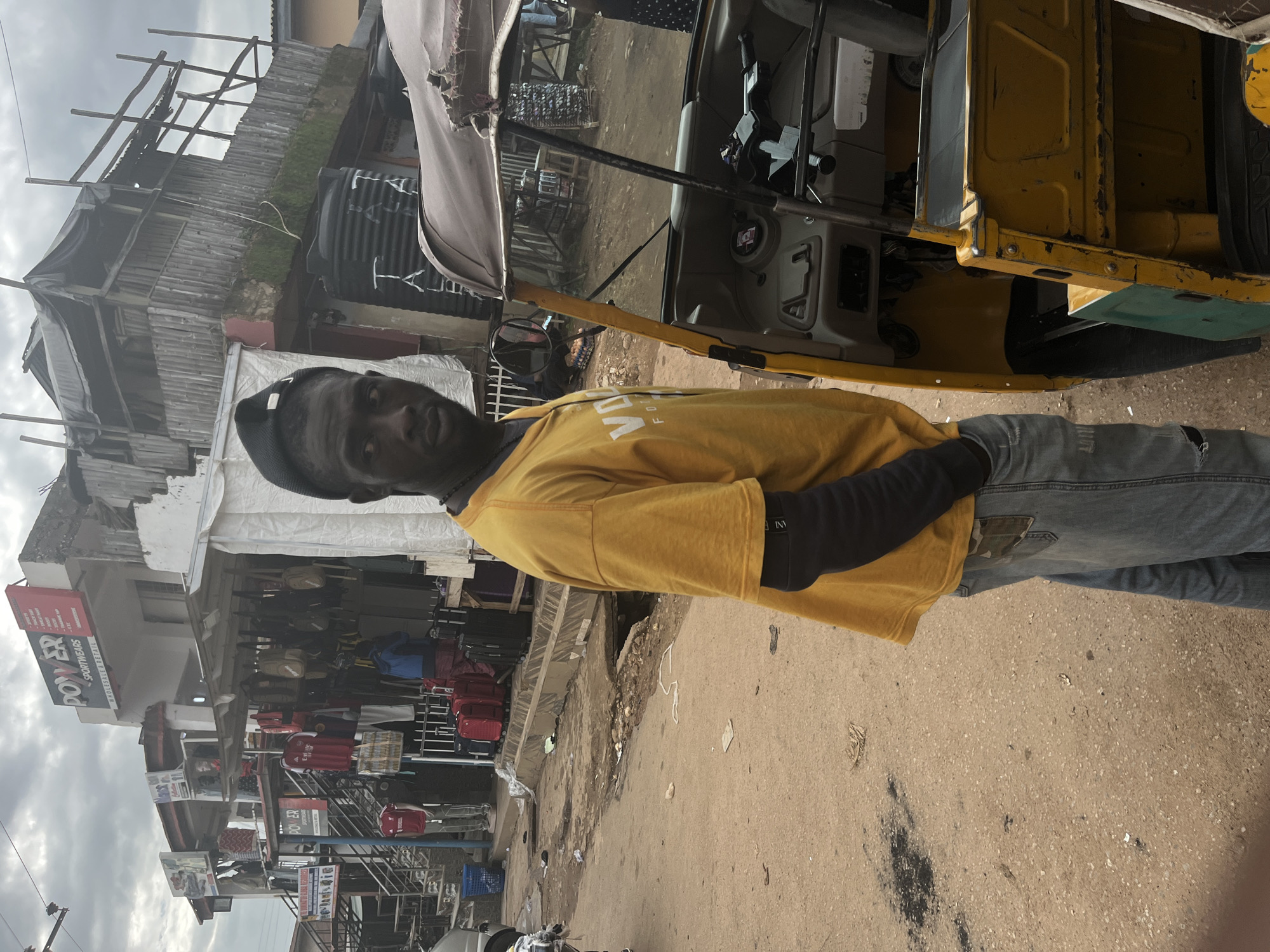 A person stands in front of a yellow motorized tricycle in a busy market area. The person is wearing a yellow t-shirt with black long sleeves underneath and blue jeans. They are looking towards the camera with a neutral expression. The tricycle appears to be open, revealing the driver's seat.
In the background, there is a row of market stalls filled with various goods, including clothing and luggage. Some of the stalls have signage, one of which reads "POWER SPORTSWEAR." The stalls are constructed with a combination of wood and metal, and some are covered with tarps. Above the stalls, there are more rudimentary structures, possibly homes or additional market extensions, made of wood and corrugated metal. A large black water tank is visible on one of the upper structures.
The ground is paved with dirt and some debris, suggesting a well-trafficked area. The sky is overcast, indicating a cloudy day. The overall scene depicts a lively market environment, likely in Jos, Nigeria, with a focus on commercial activity and everyday life. The image is oriented vertically.