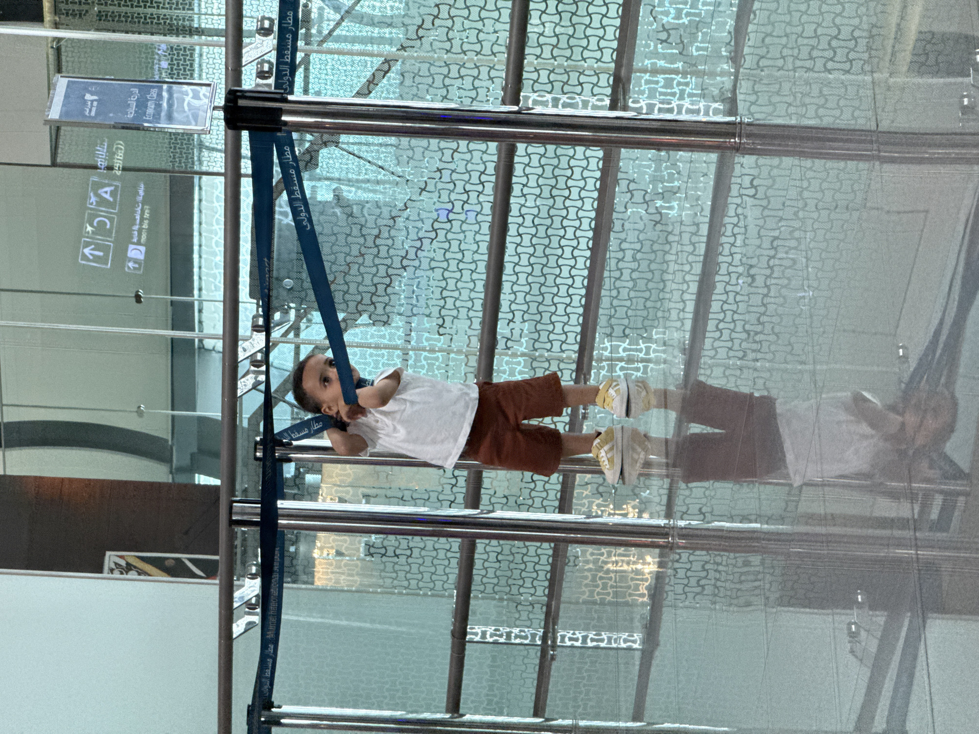 A young child wearing a white t-shirt and brown shorts stands in what appears to be an airport terminal. The child is leaning against a blue rope barrier with Arabic text on it, which separates the waiting area from a pathway. The child's reflection is visible on the highly polished floor. In the background, there are glass partitions with a patterned design, and signs indicating "Economy Class" and directional arrows are visible on a reflective surface. The overall environment suggests a modern, indoor public space, likely Muscat International Airport, located in Seeb Province, Oman.