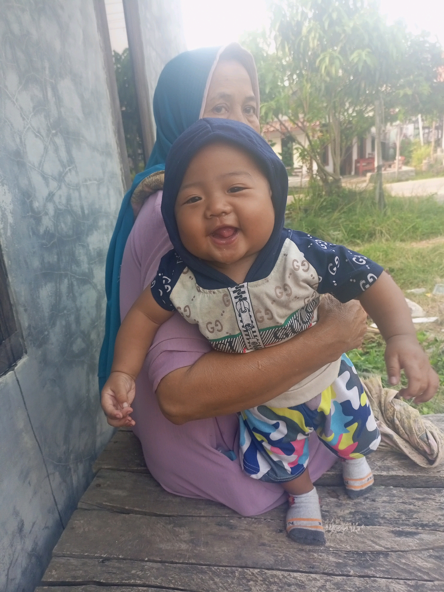 In a medium shot, a woman wearing a light blue hijab and a lavender shirt holds a baby in her arms. The baby, wearing a dark blue hooded shirt with a beige and patterned design and colorful camouflage shorts with white socks, is looking forward with its mouth open and a wide smile. The woman's left arm is around the baby's back, while her right arm supports the baby's legs. They are sitting on a weathered wooden bench. To the left of the bench is a textured, light gray wall. In the background, there are green trees and what appears to be houses, suggesting a residential outdoor setting in Jawilan, Indonesia. The lighting suggests it is daytime, possibly with bright sunlight causing some overexposure in the upper right portion of the image.
