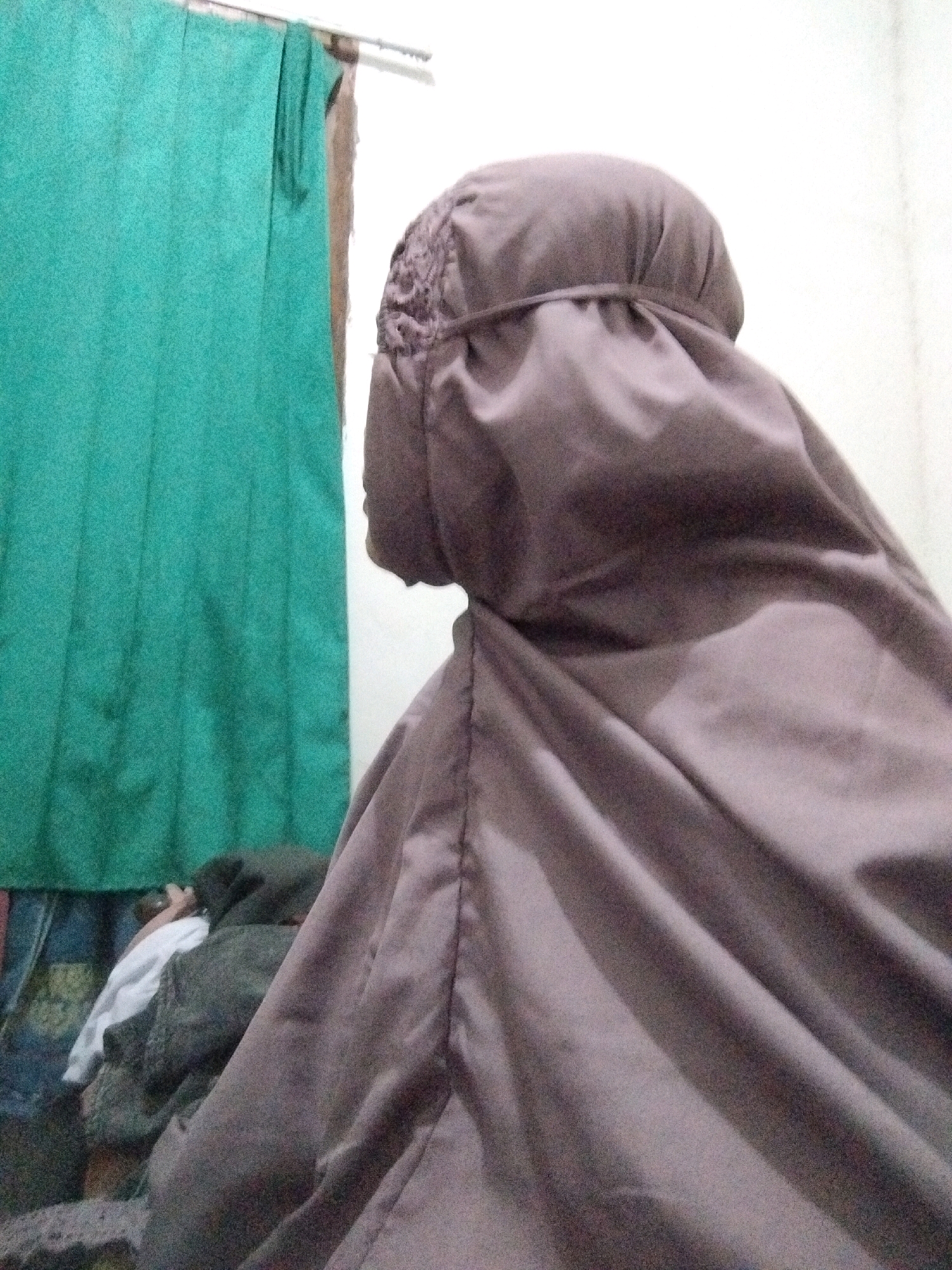 The image shows a person wearing a brown hijab, with a green curtain in the background. The setting appears to be indoors, possibly a bedroom, with some clothing and blankets visible in the lower left portion of the frame. The image is not specific to Buntalan, Indonesia, and does not provide enough visual information to determine a precise location within a city.